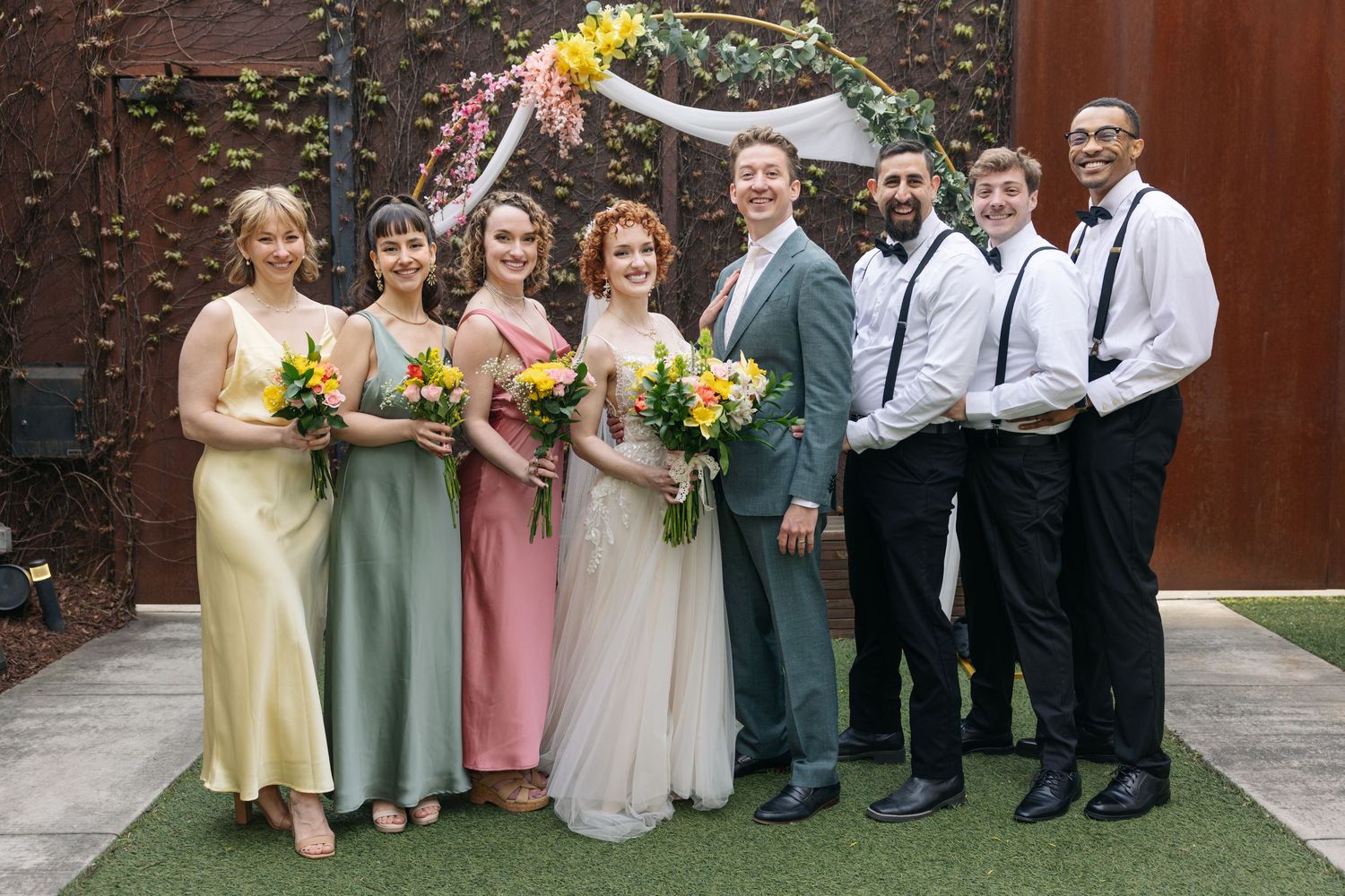 Wedding party poses formally with bride and groom flanked by bridesmaids and groomsmen outdoors.
