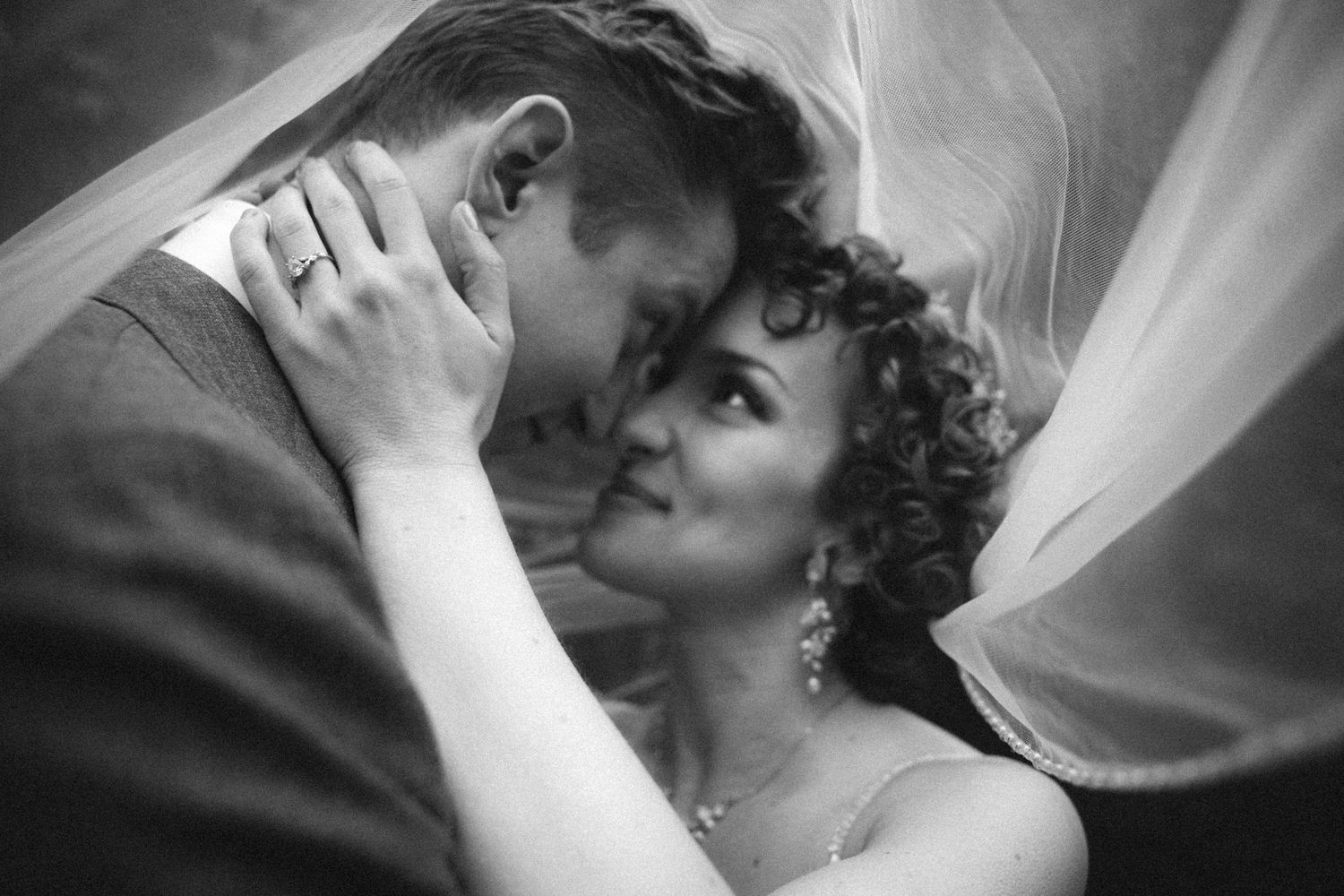 Black and white romantic portrait of bride and groom sharing intimate moment beneath flowing veil.