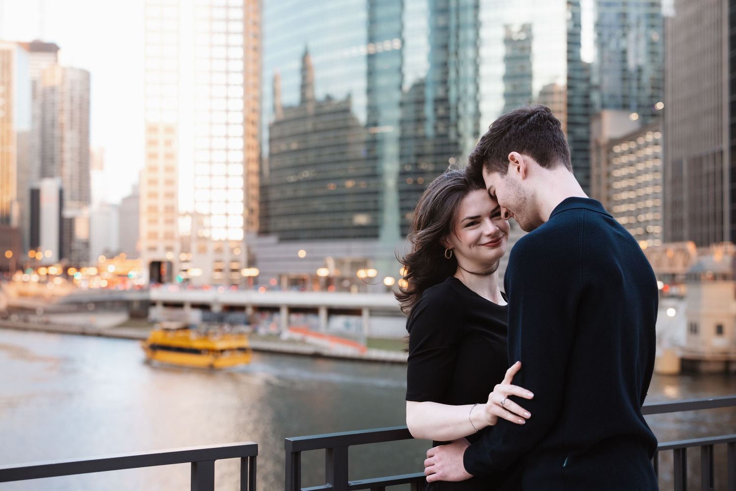 A smiling woman embraces her partner on a riverside bridge with Chicago skyline at golden hour.