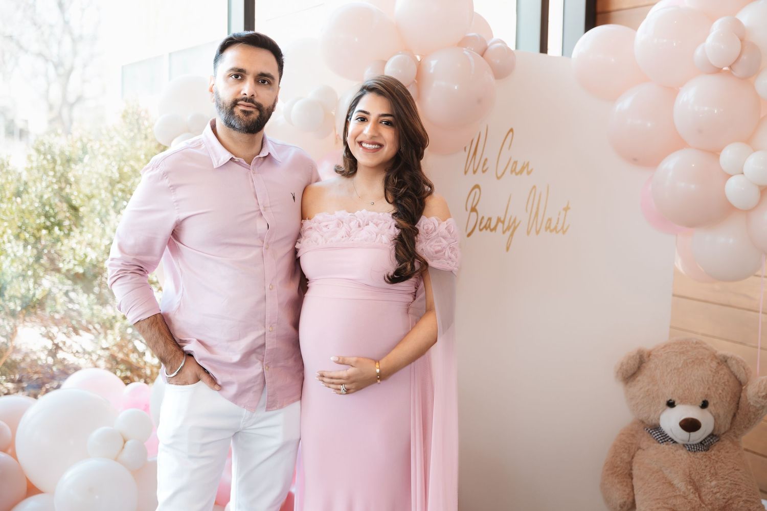 South Asian expecting couple in coordinating pink outfits pose at We Can Bearly Wait baby shower.