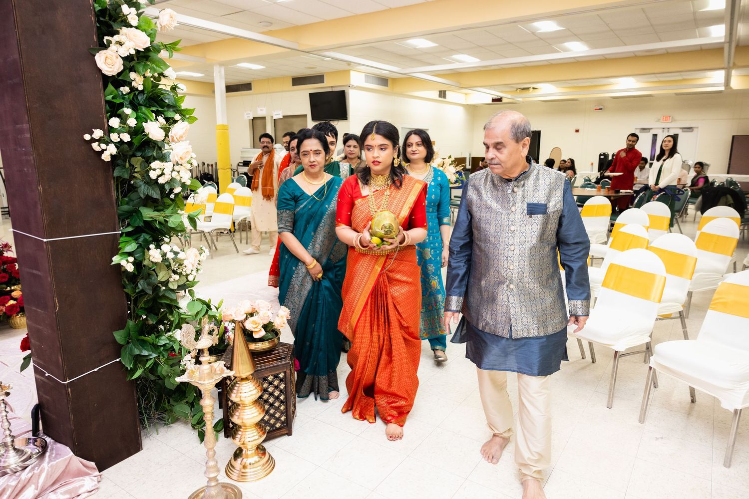 Indian bride in red saree walking down the aisle with family members during traditional South Indian wedding ceremony.