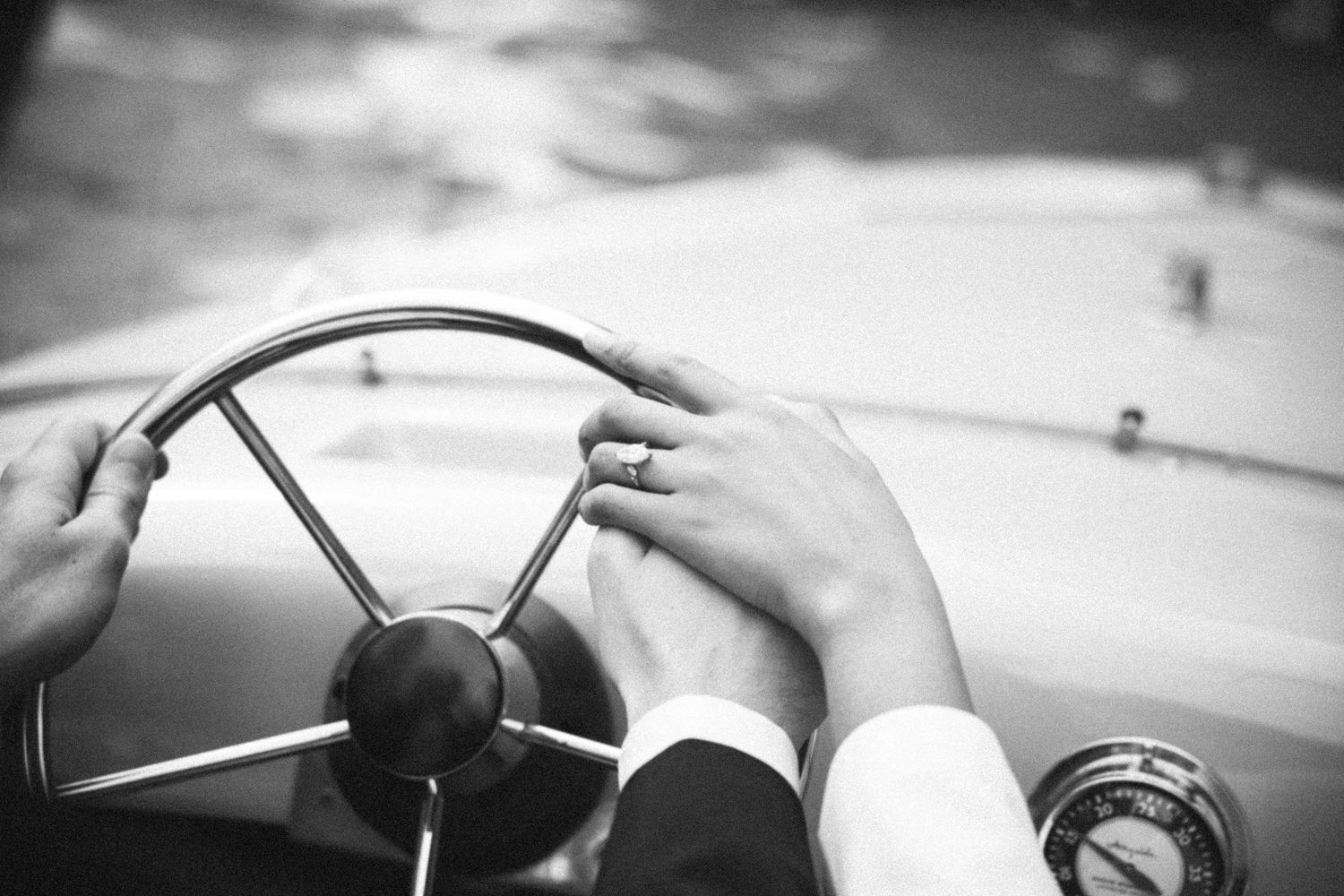 Black and white photo of two hands on a vintage car steering wheel, one wearing a wedding ring.