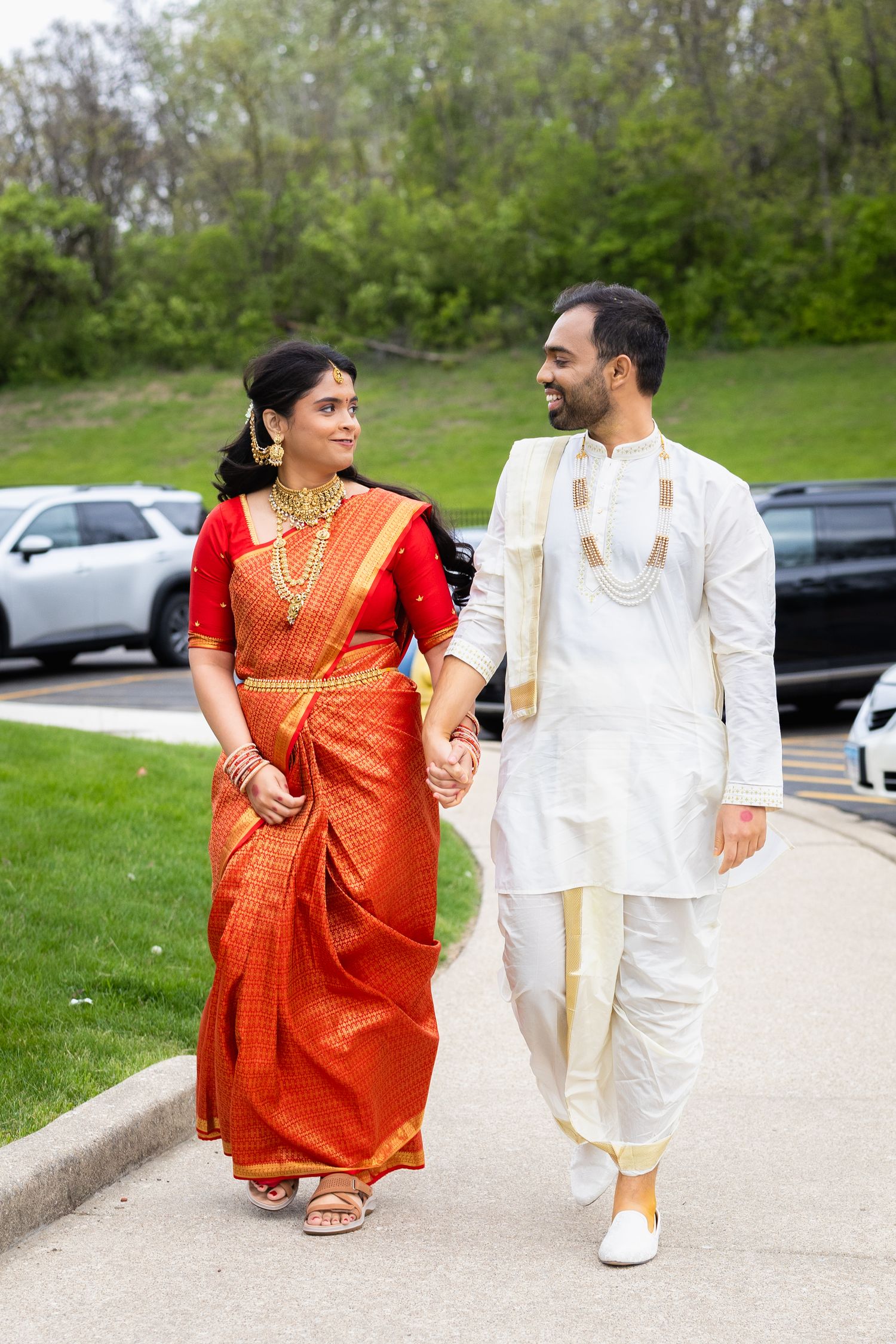 Indian couple in traditional wedding attire walking hand in hand, bride in red saree and groom in white kurta.