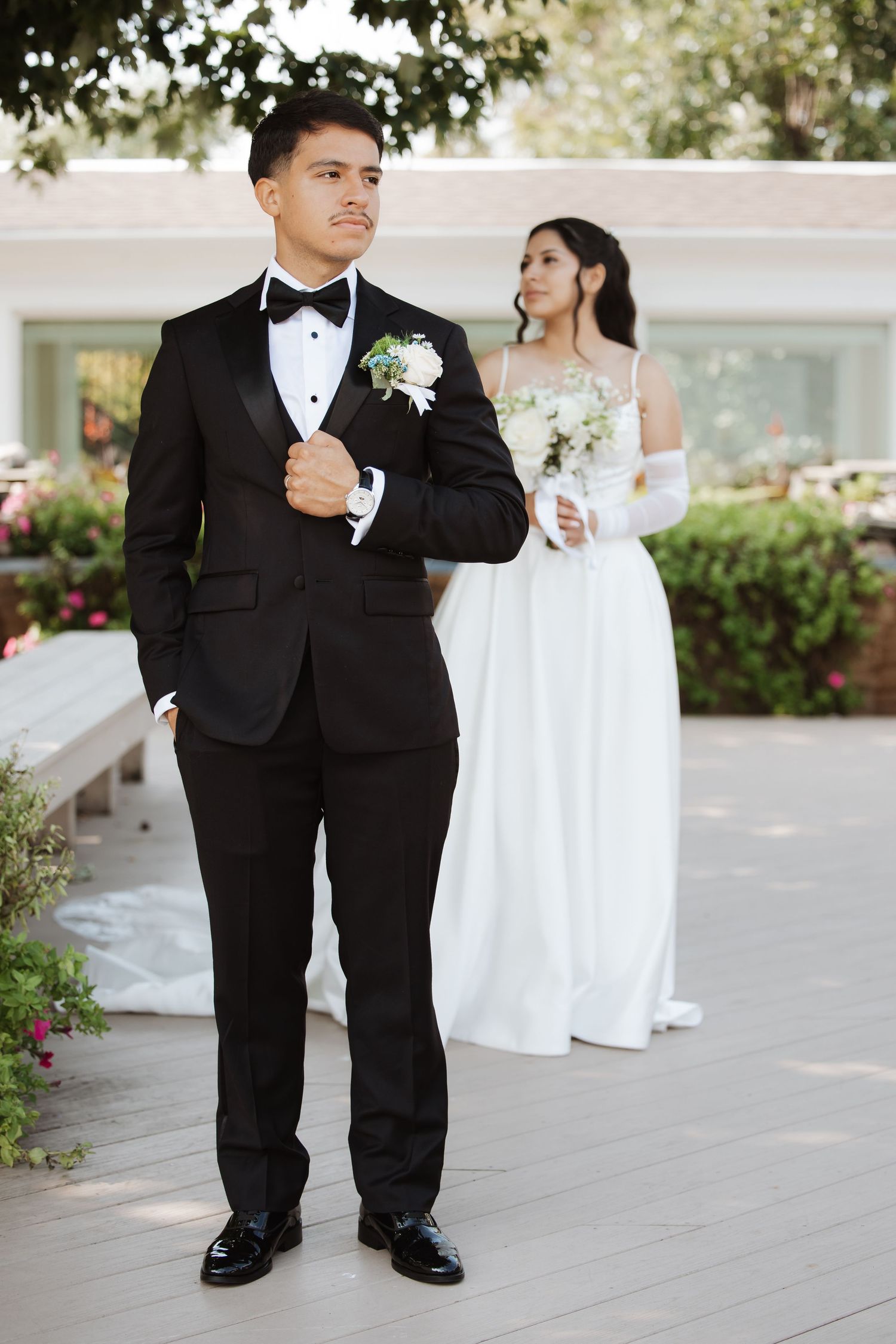 Groom in black tuxedo stands confidently as bride in white dress holds bouquet behind him.