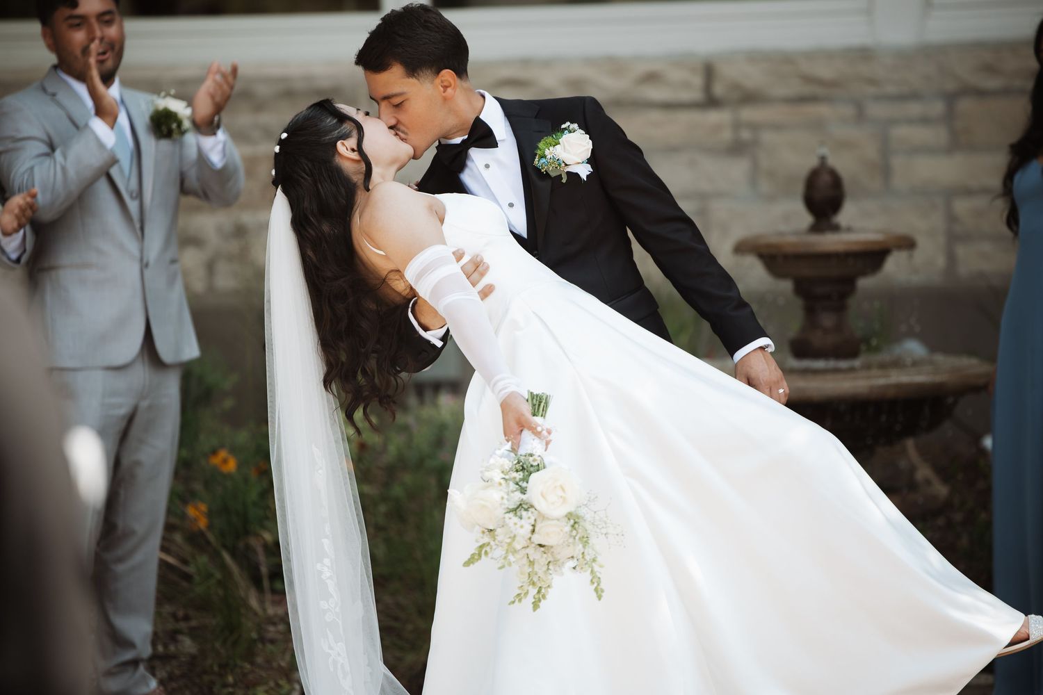 Groom dips bride in white gown for romantic kiss near stone fountain during outdoor wedding.