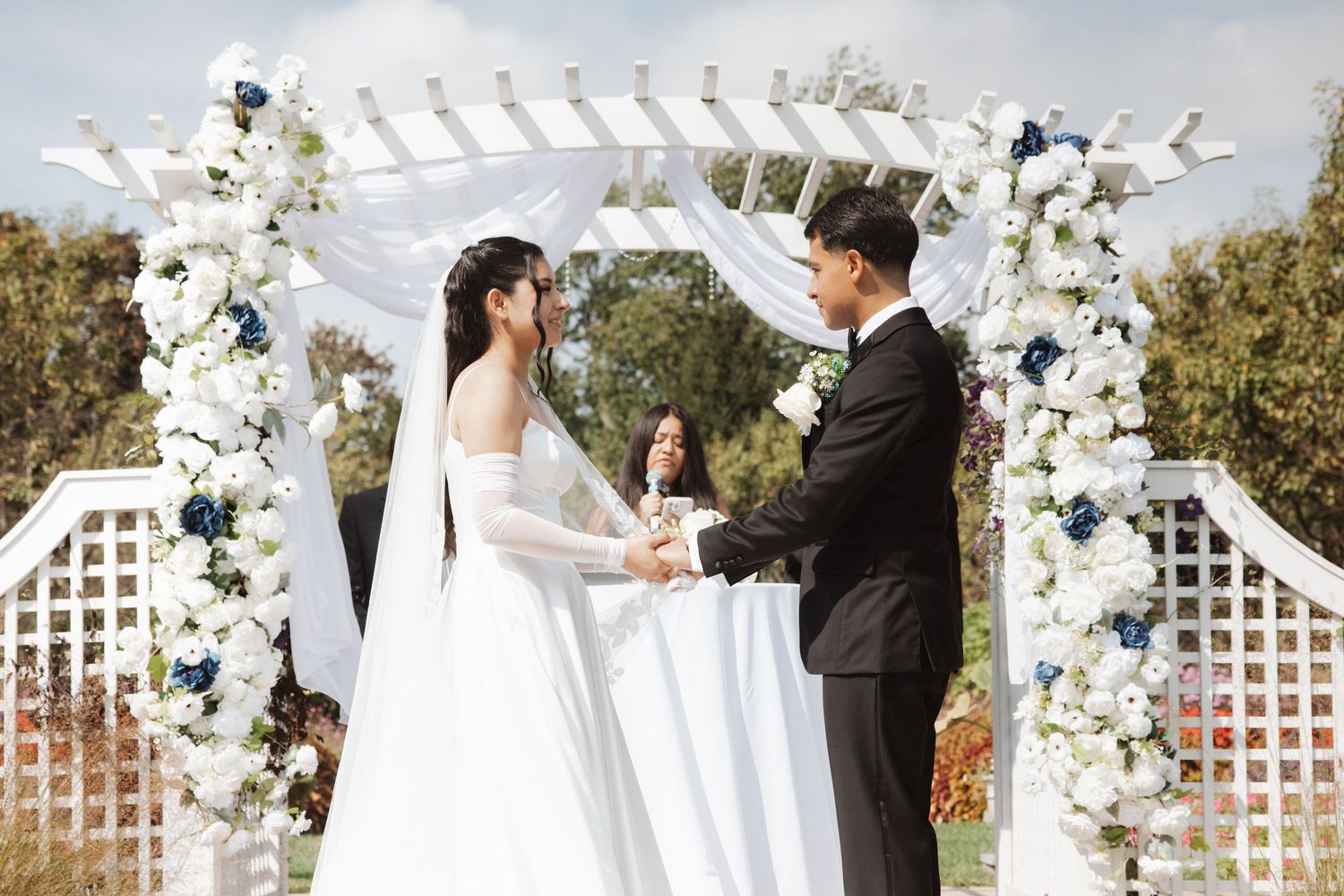 Bride and groom hold hands under white floral arch at sunny outdoor wedding ceremony.