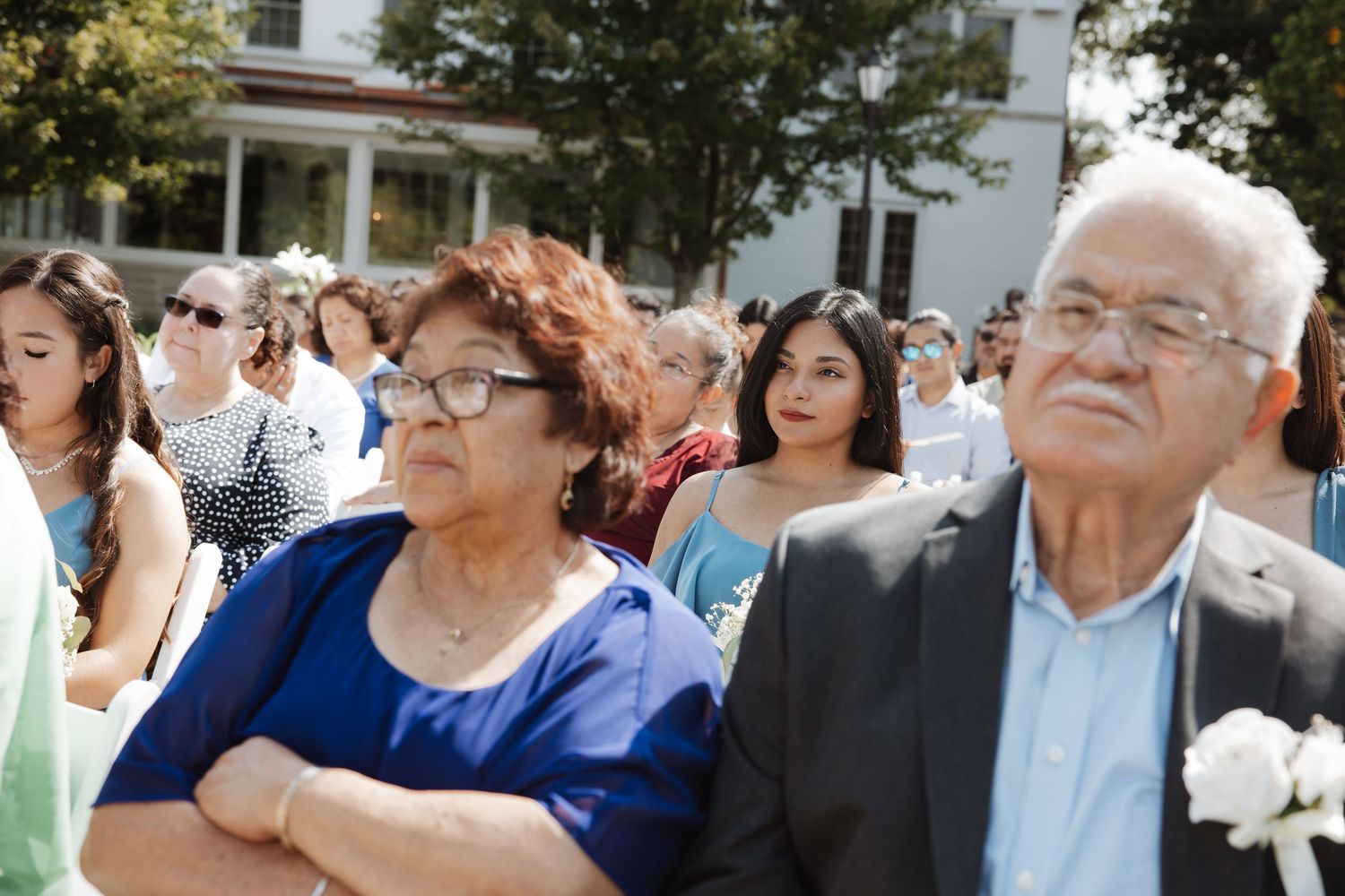 An elderly couple dressed formally watches an outdoor wedding ceremony surrounded by other guests.
