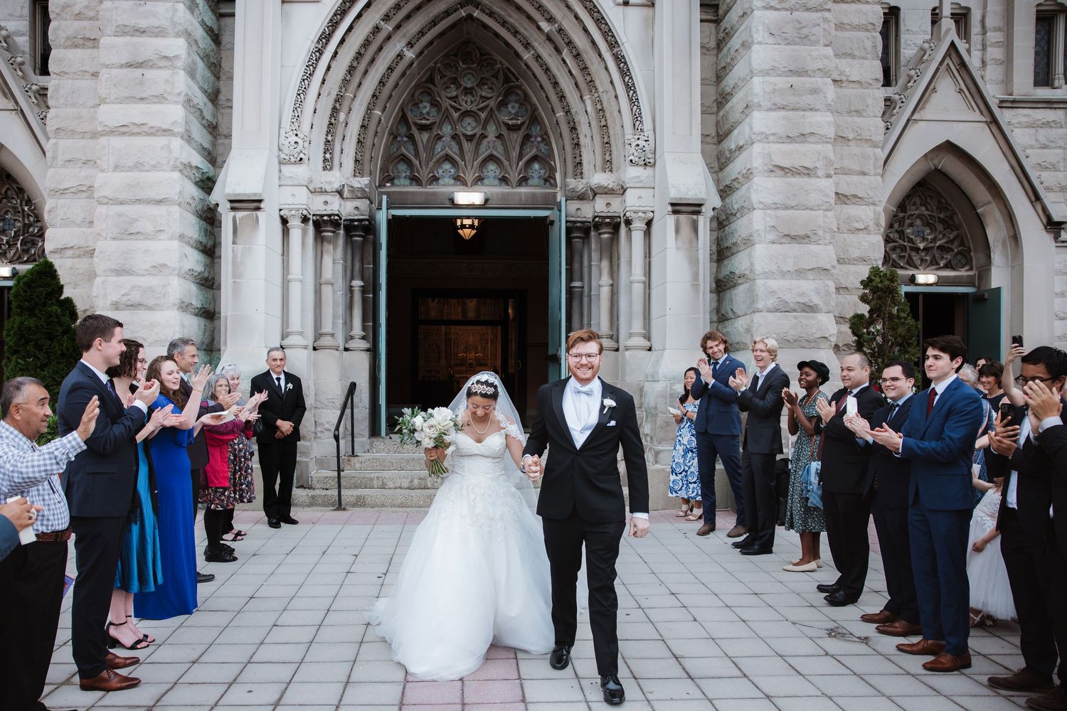 Bride and groom exit gothic church doors as wedding guests applaud and celebrate their marriage ceremony.
