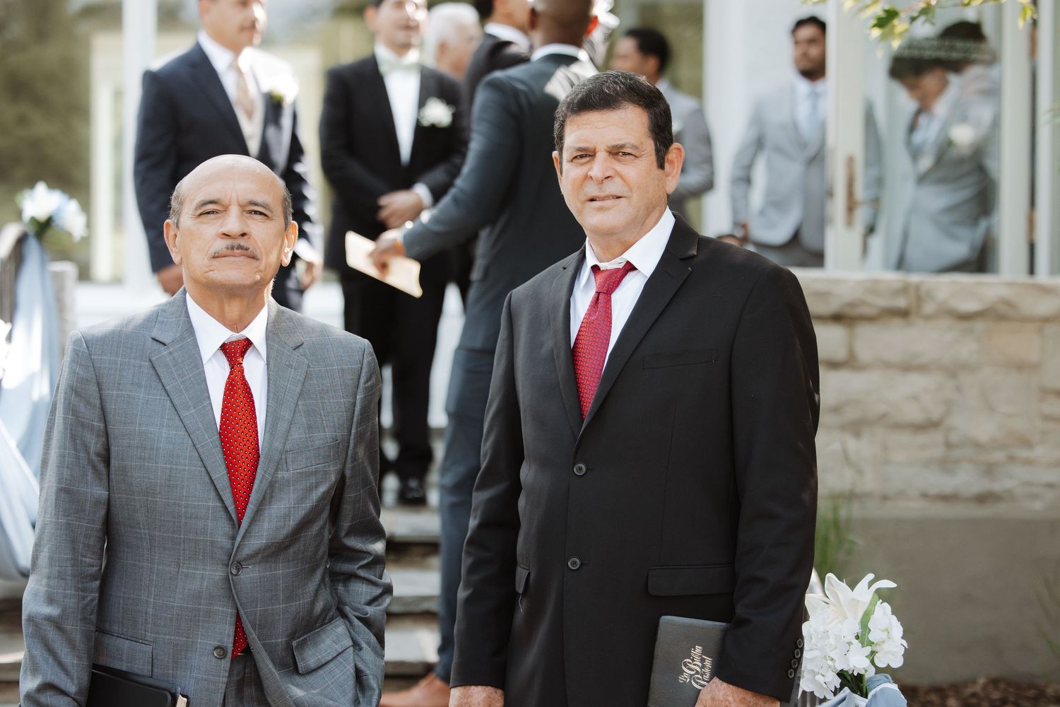 Two men in black suits and red ties smile outdoors at a sunny wedding celebration.