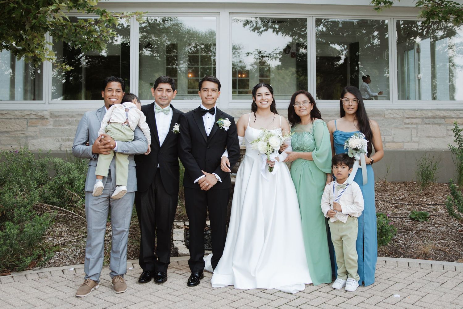 Wedding party group photo featuring bride in white gown, groom in tuxedo, and family outside venue.