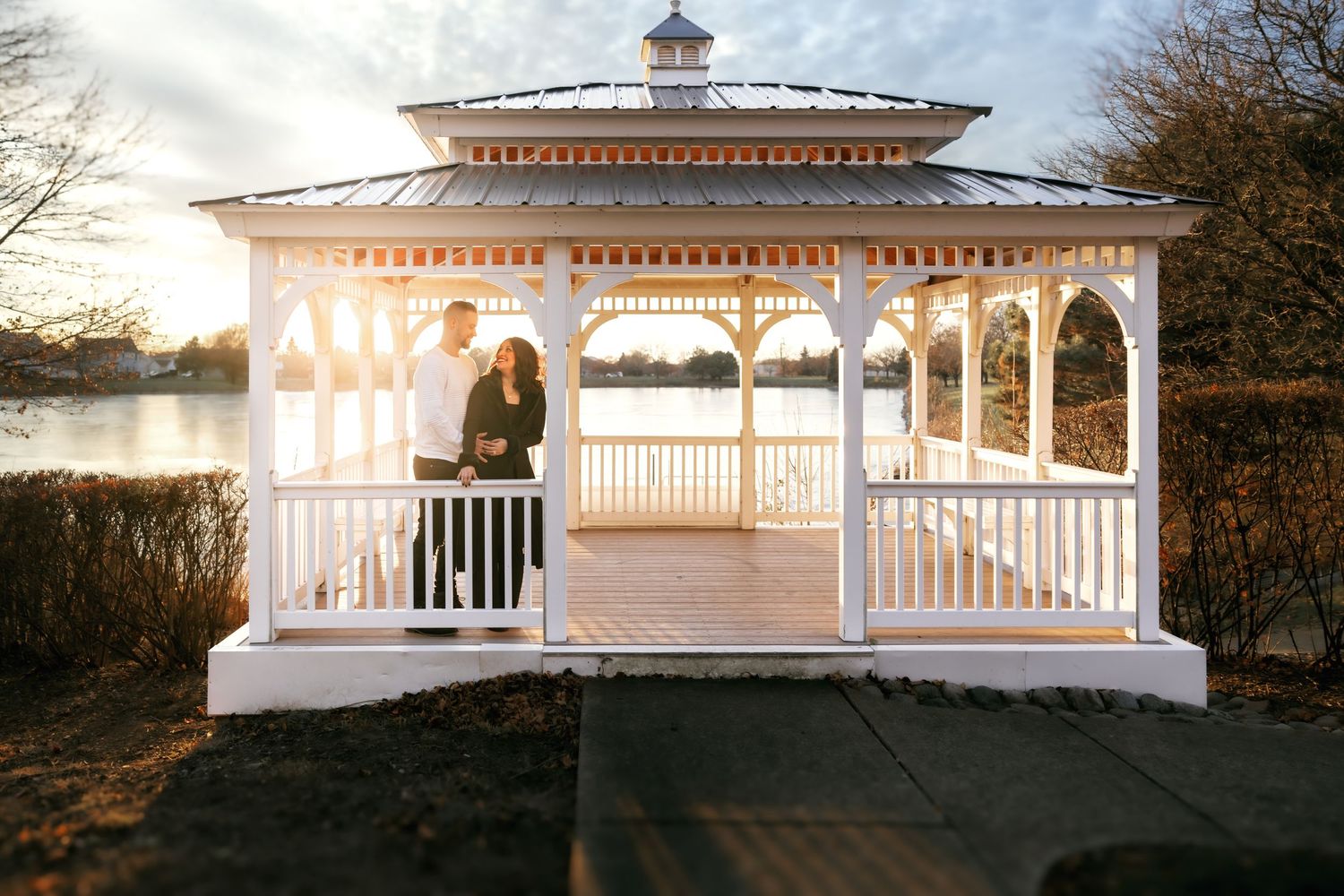 Couple standing together in a charming white gazebo at golden hour with a peaceful lake backdrop.