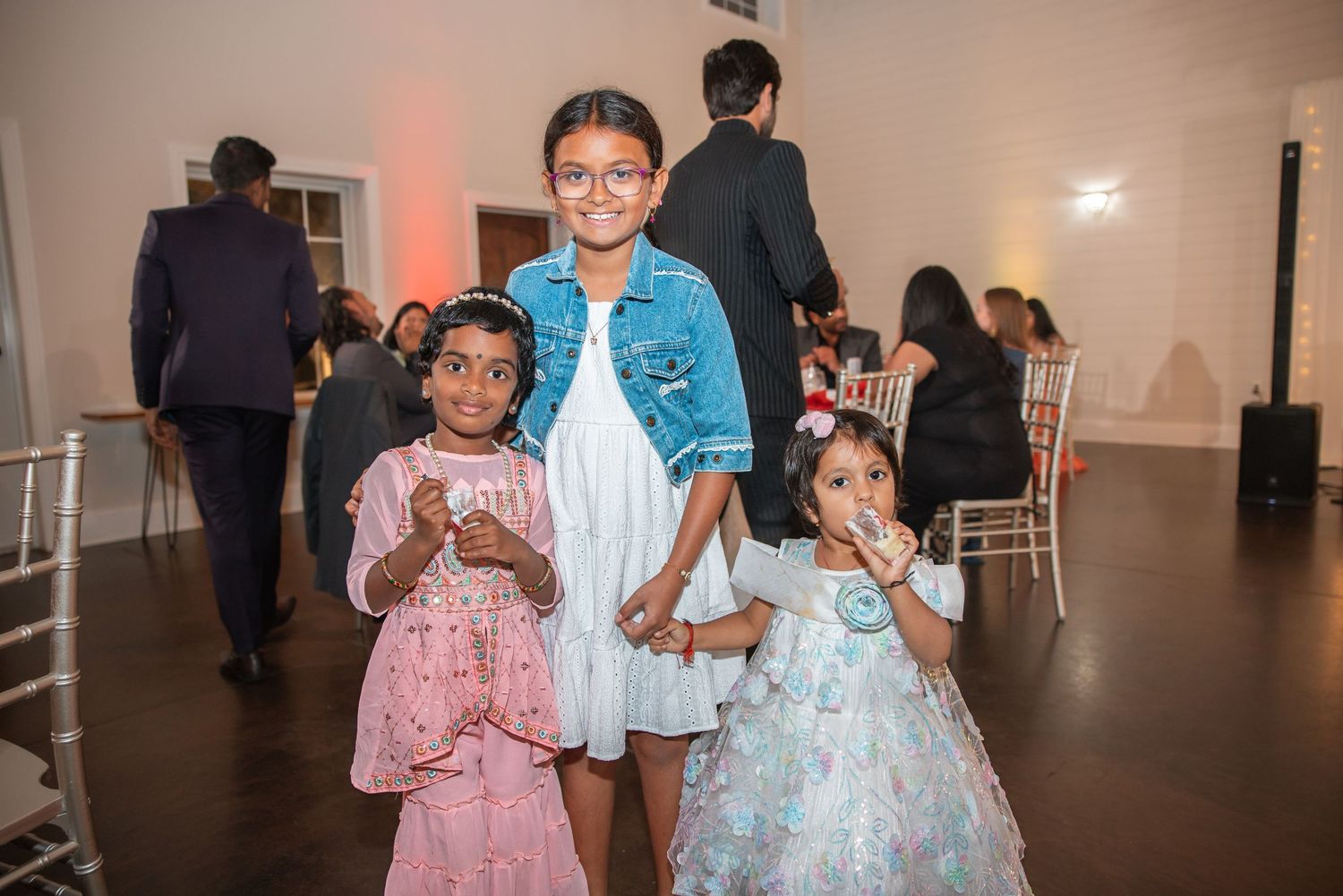 Three young Indian girls in colorful traditional outfits smiling together at a festive wedding celebration event.