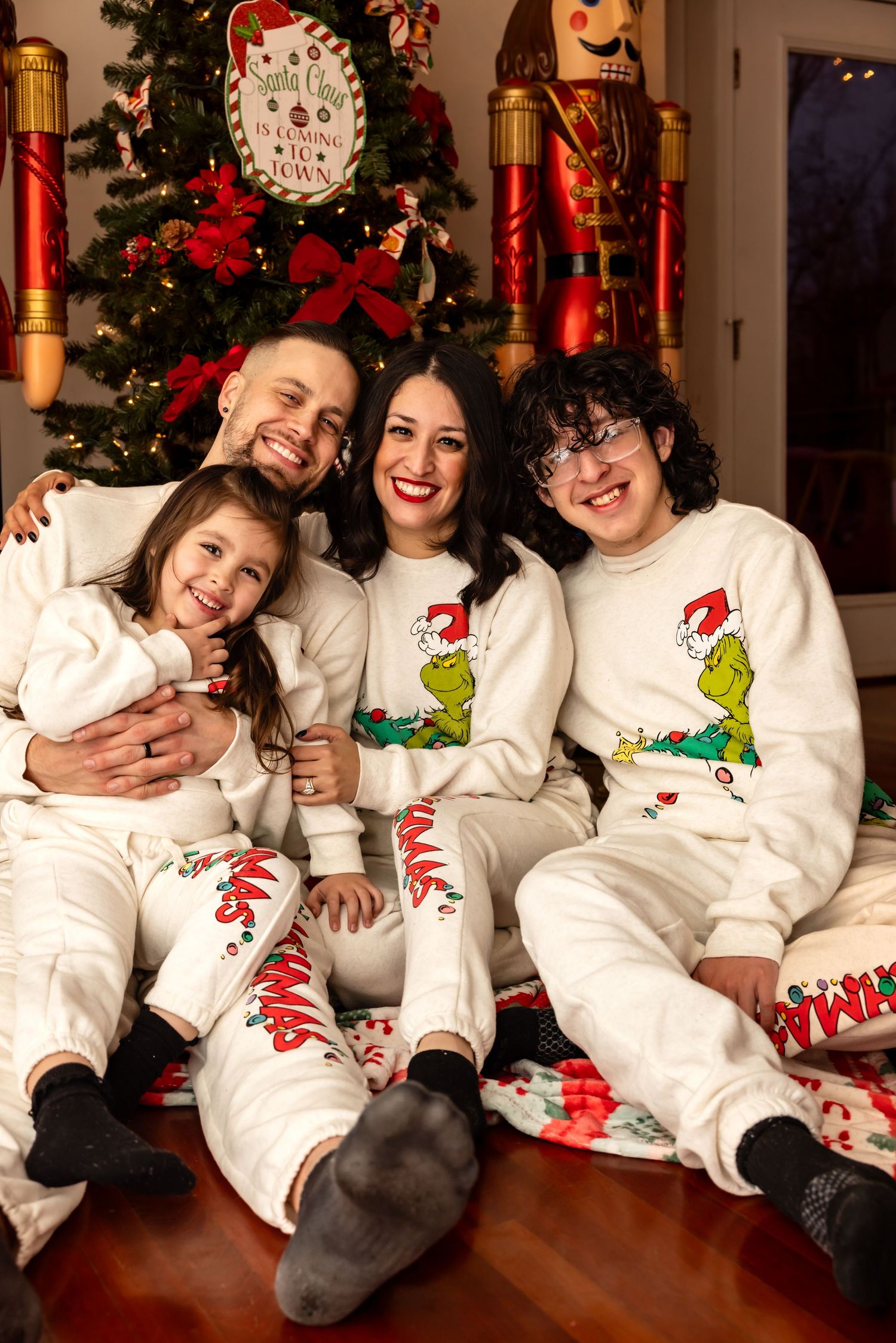 Family in matching Grinch pajamas sitting on floor by decorated Christmas tree.