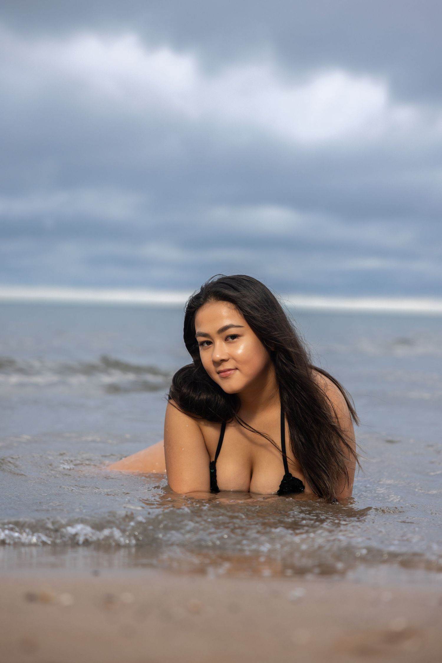 Woman in black bikini lying in shallow beach waves, smiling at camera with cloudy sky and ocean backdrop.