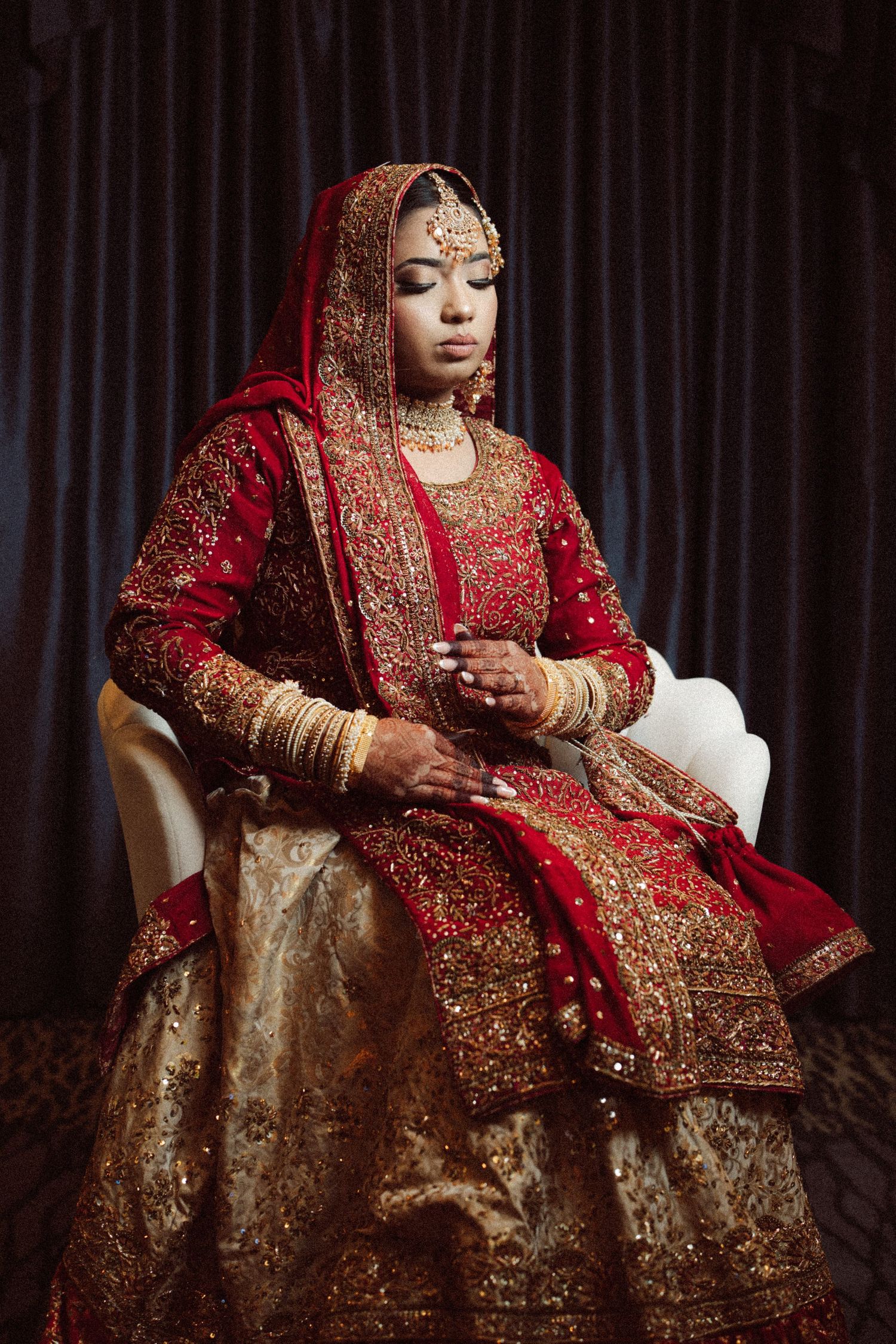 A bride in traditional red and gold South Asian bridal wear sits gracefully in a white chair.