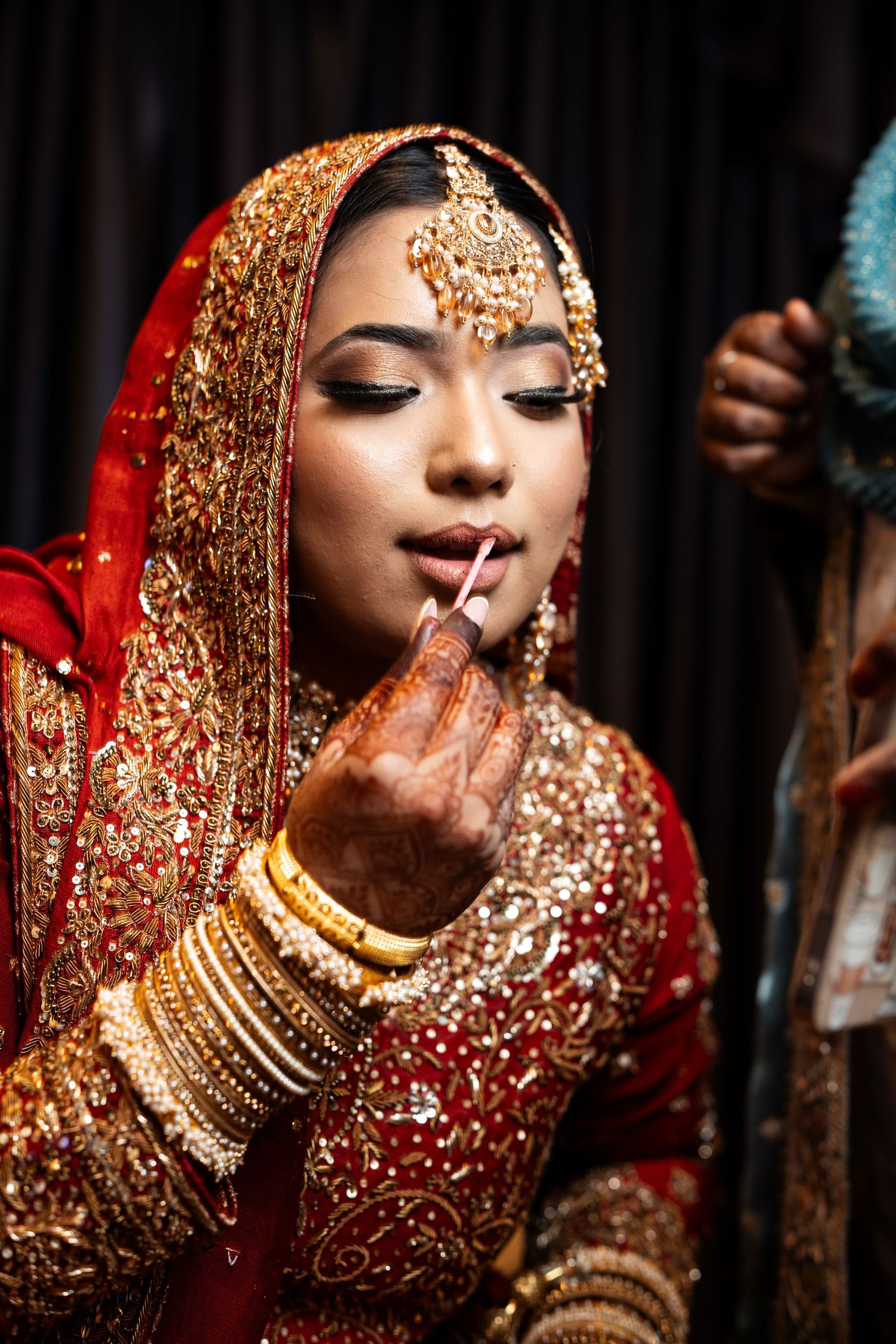 Indian bride in red bridal lehenga applying lipstick with mehndi-adorned hands and gold jewelry.