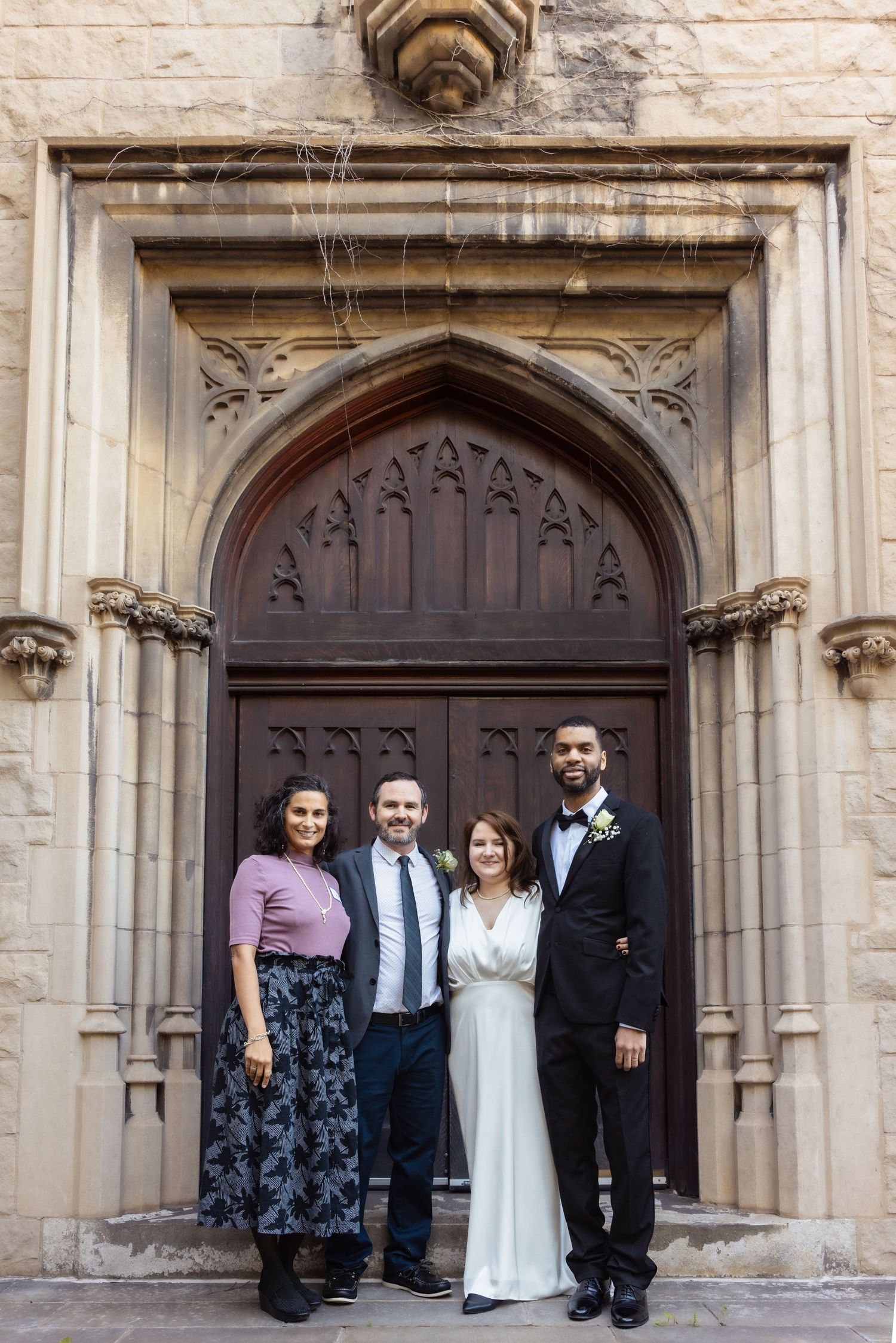 Two couples pose in formal attire before a Gothic stone church doorway.