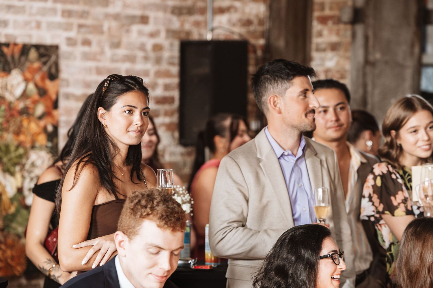 Guests in smart casual attire socializing and watching speeches at an indoor event or wedding reception.