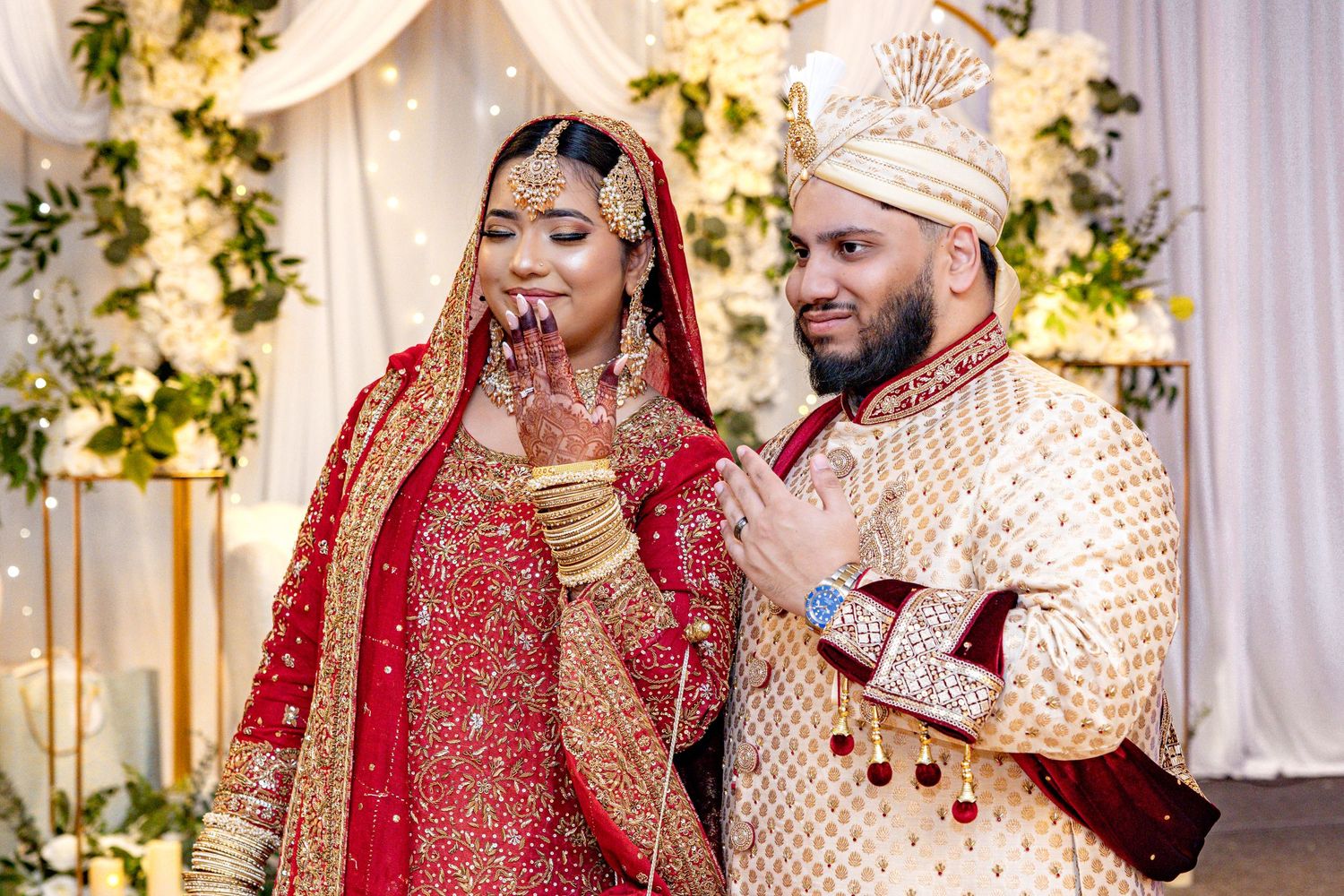 Indian bride in red bridal lehenga and groom in gold sherwani posing at wedding ceremony.