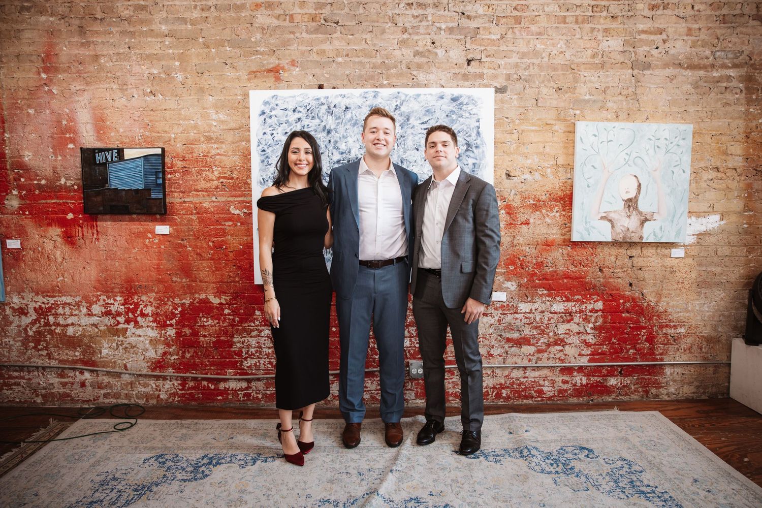 Three professionals smiling together in front of large artwork at an art gallery reception.