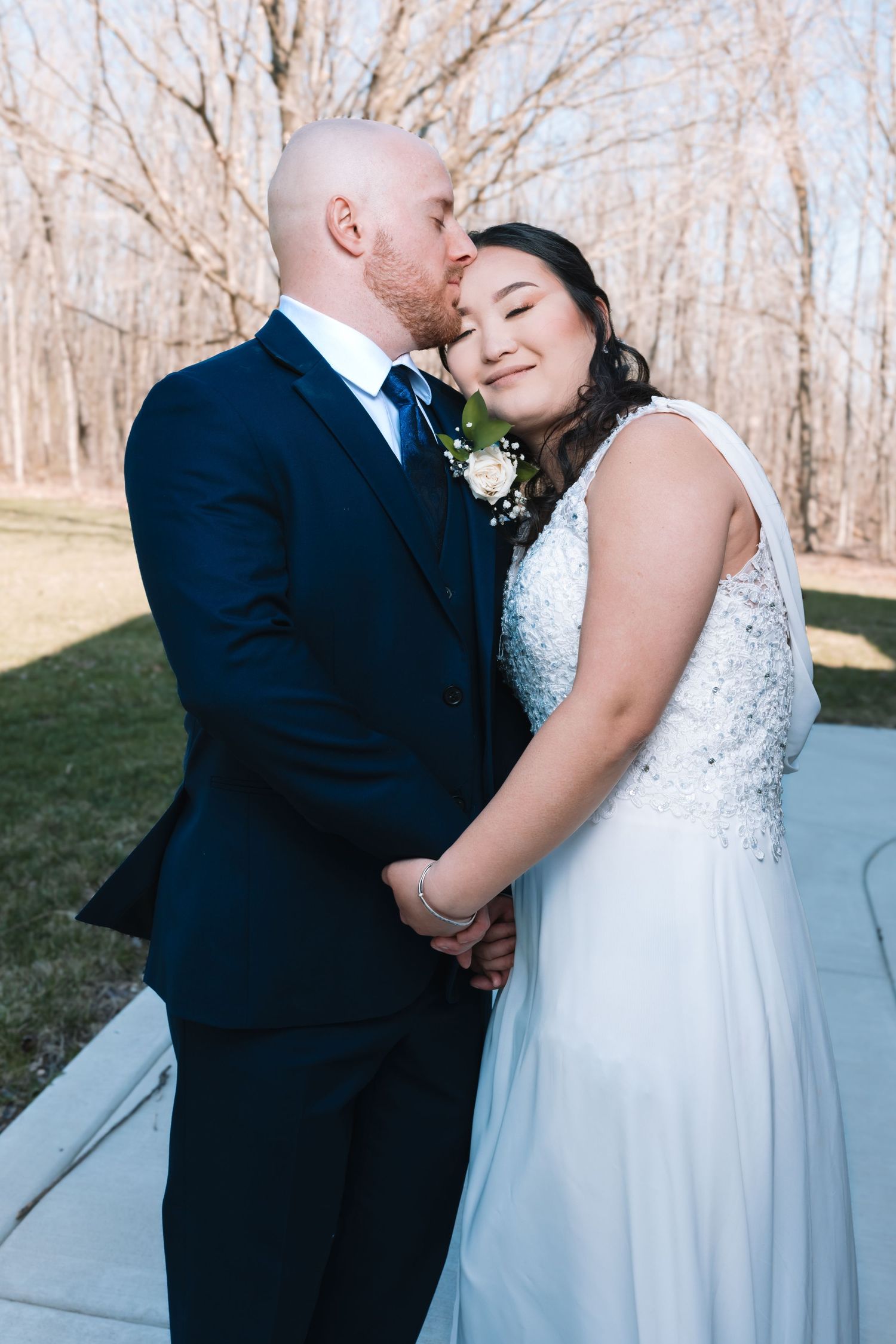 Groom in navy suit kisses bride's cheek as she smiles with eyes closed, holding hands outdoors.