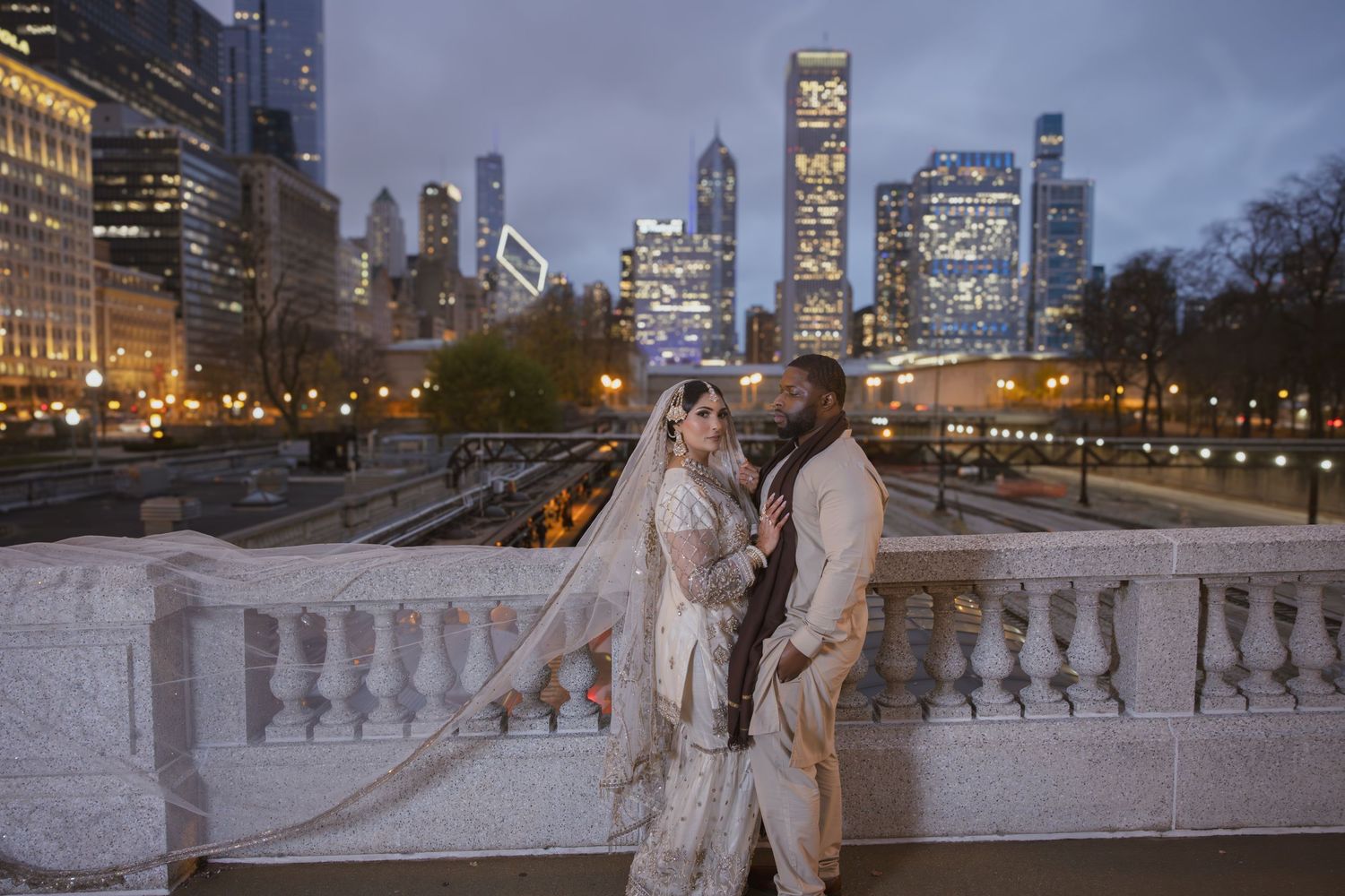 Wedding couple in traditional South Asian attire embrace on bridge with Chicago skyline at night.