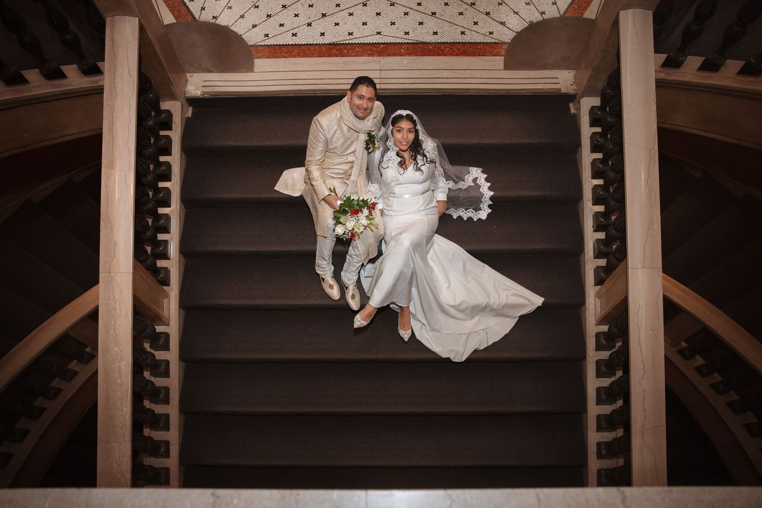 Overhead shot of bride and groom sitting on wooden spiral staircase, white wedding dress flowing elegantly.