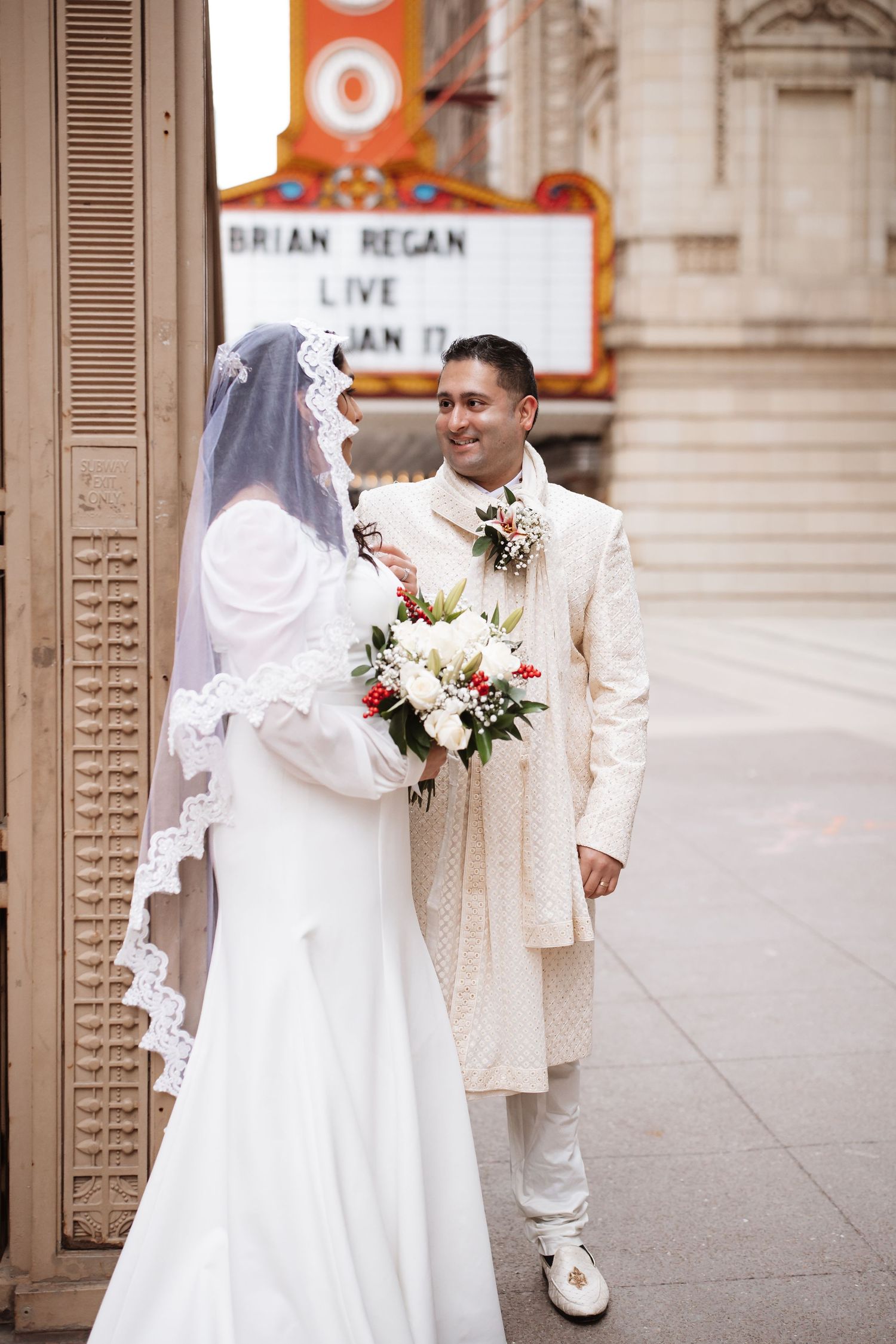 Groom in cream sherwani smiles at bride holding bouquet outside Chicago Theatre marquee.