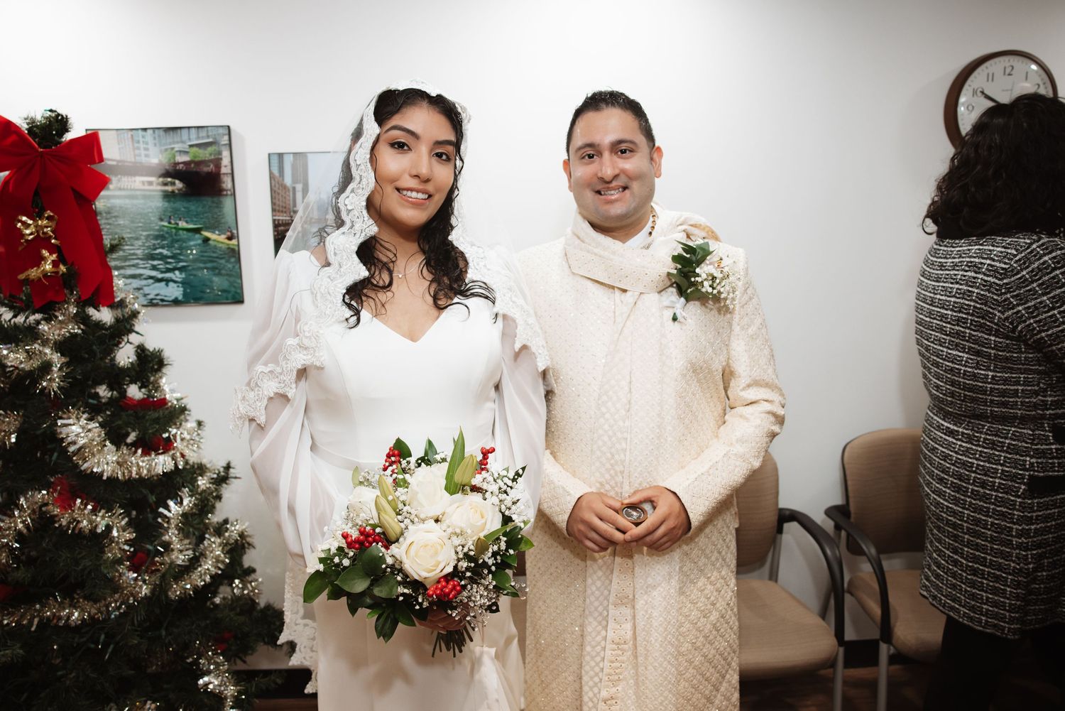 Bride in white gown holds red and white bouquet while standing with groom near decorated Christmas tree.