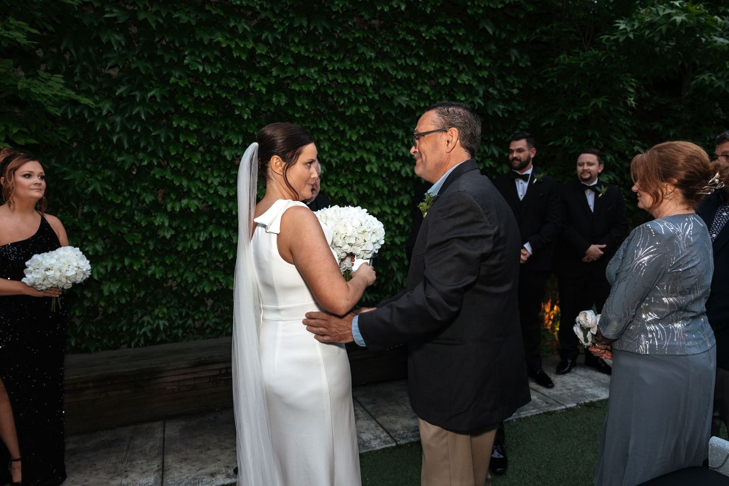 Bride in white gown holds bouquet while speaking with a man during an outdoor wedding ceremony.