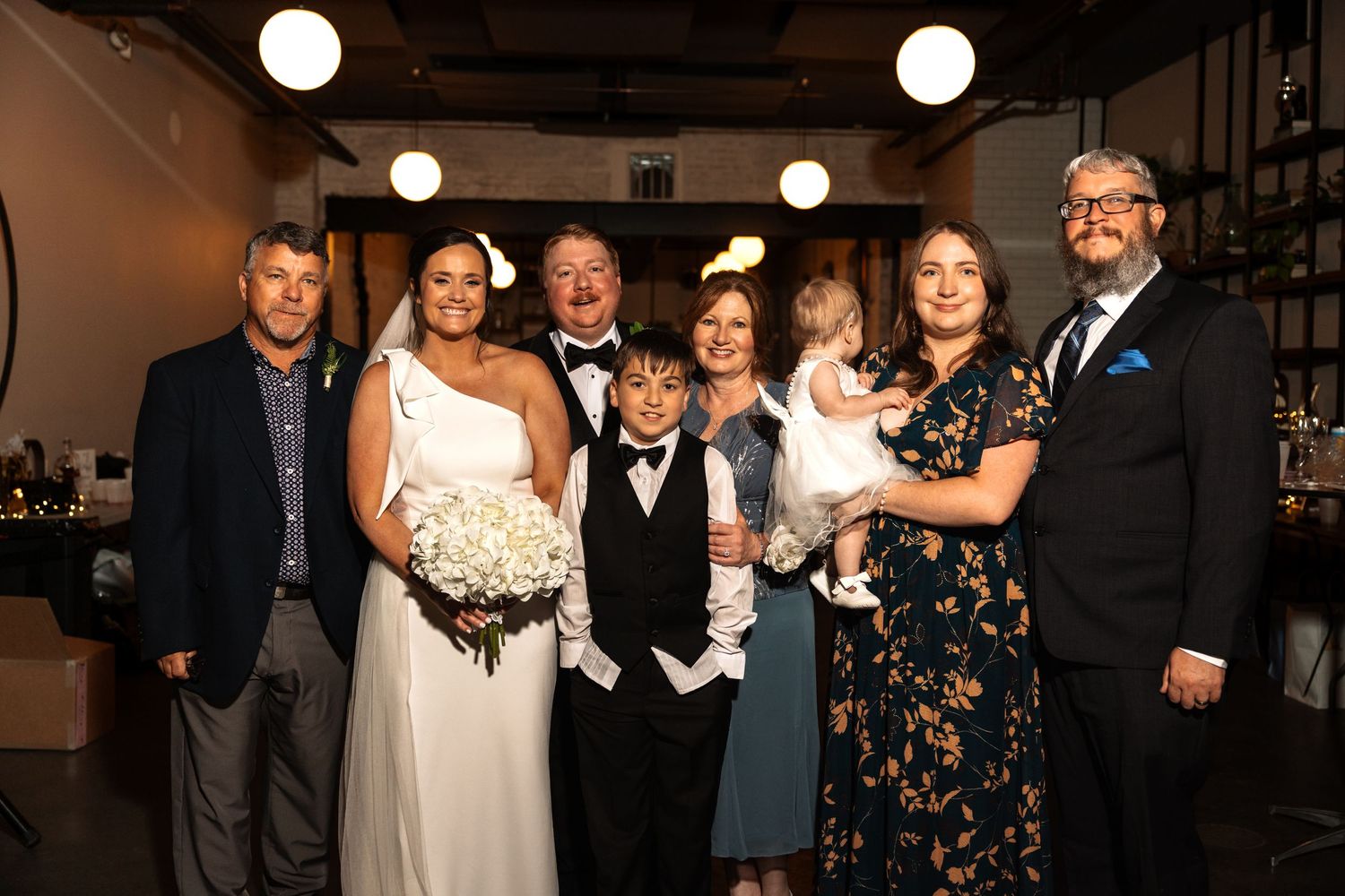 Bride and groom celebrate with seven family members including children at warmly lit indoor wedding reception.