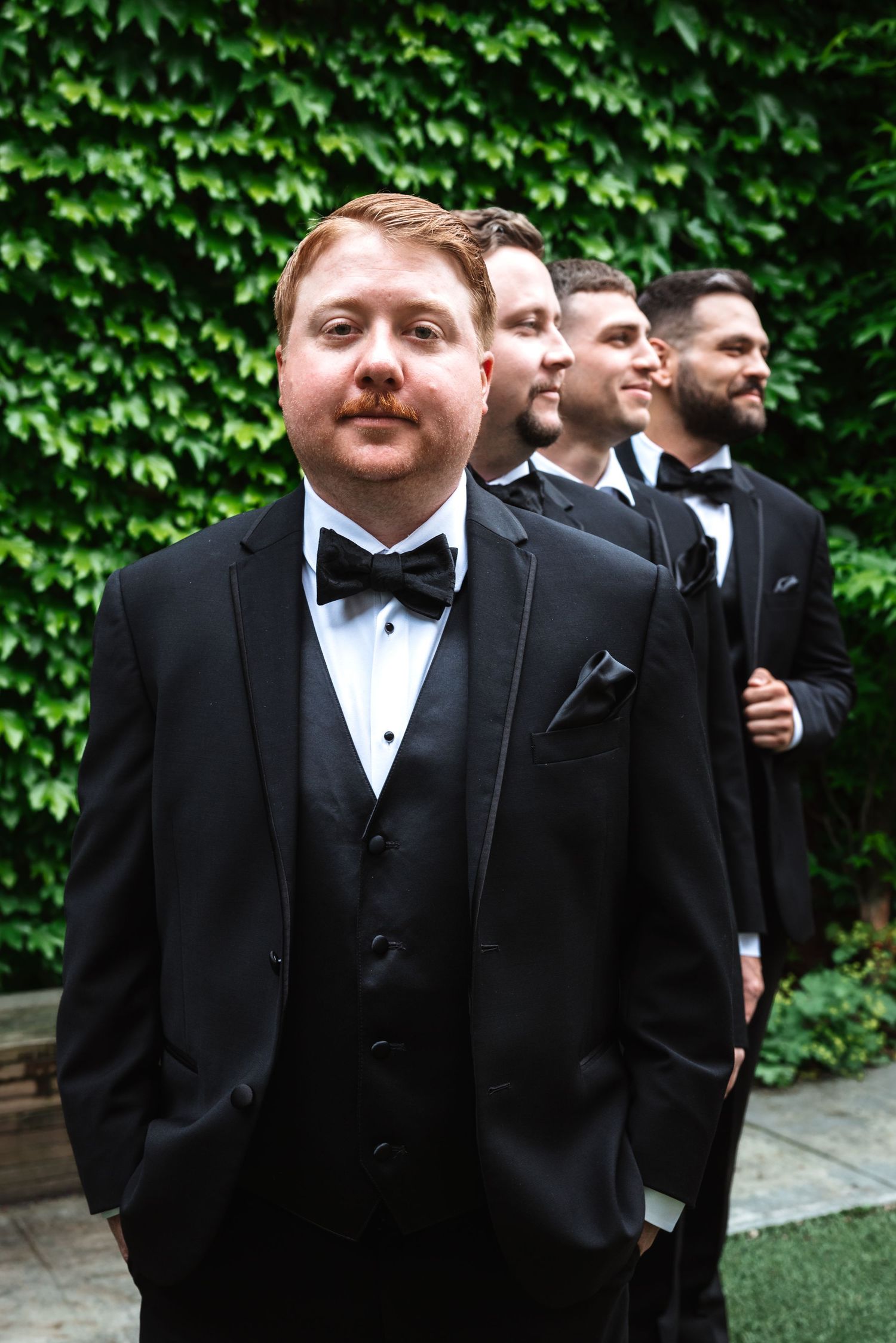 Groom in black tuxedo with bow tie stands confidently with groomsmen lined behind him against ivy wall.