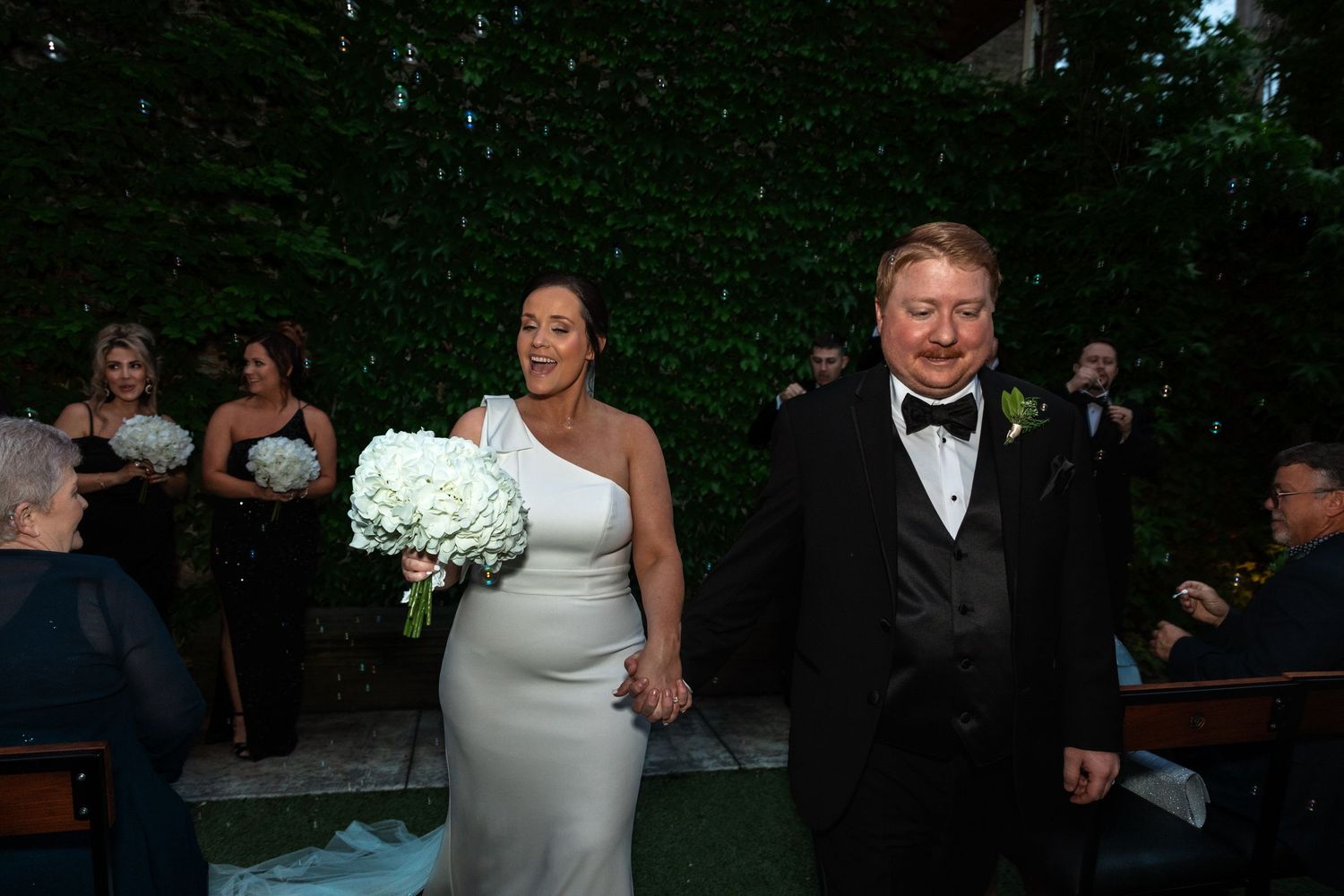 Newlyweds walk hand-in-hand down the aisle, bride holding white floral bouquet at elegant outdoor wedding ceremony.