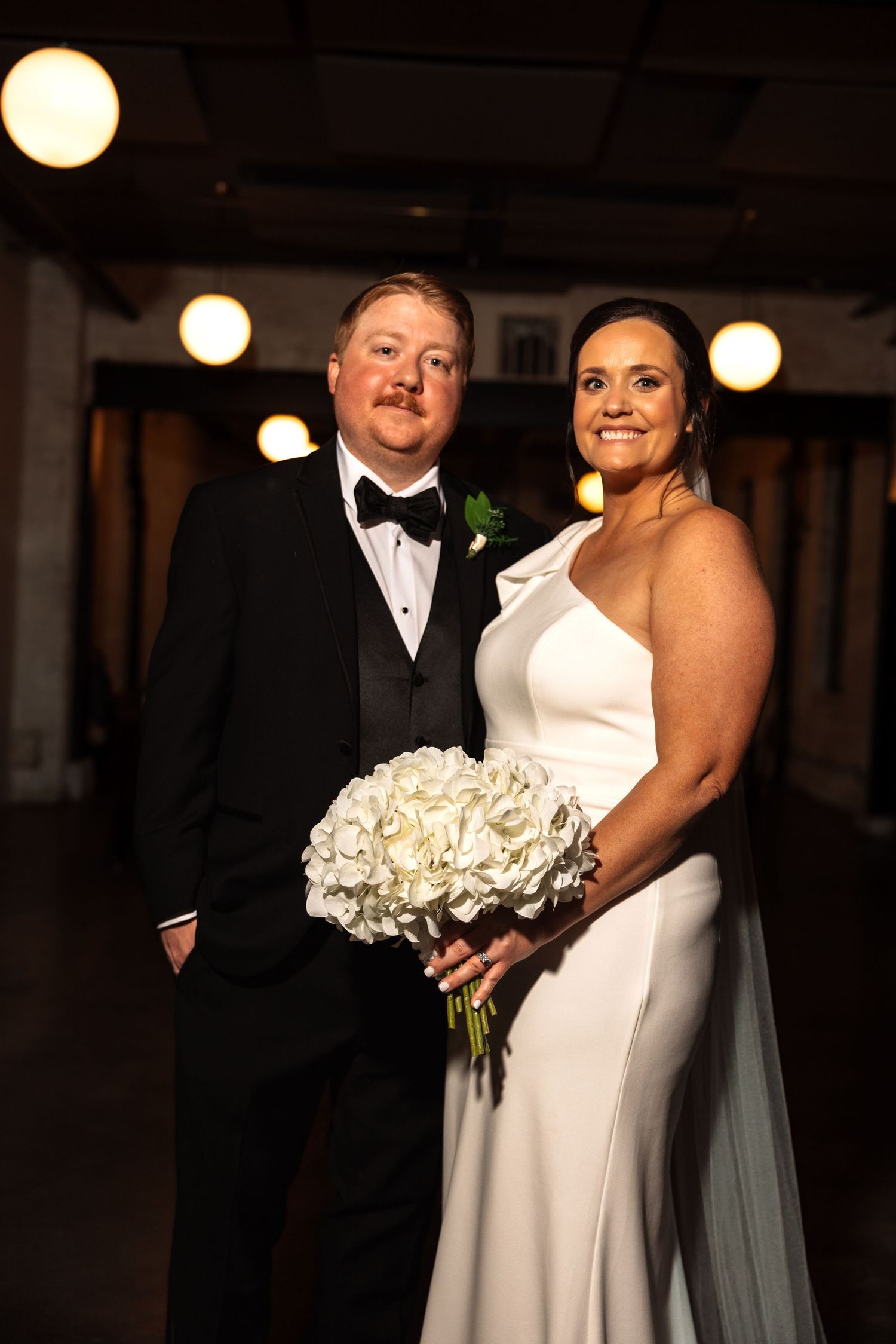 Smiling couple on wedding day, groom in tuxedo, bride in white gown holding hydrangea bouquet.
