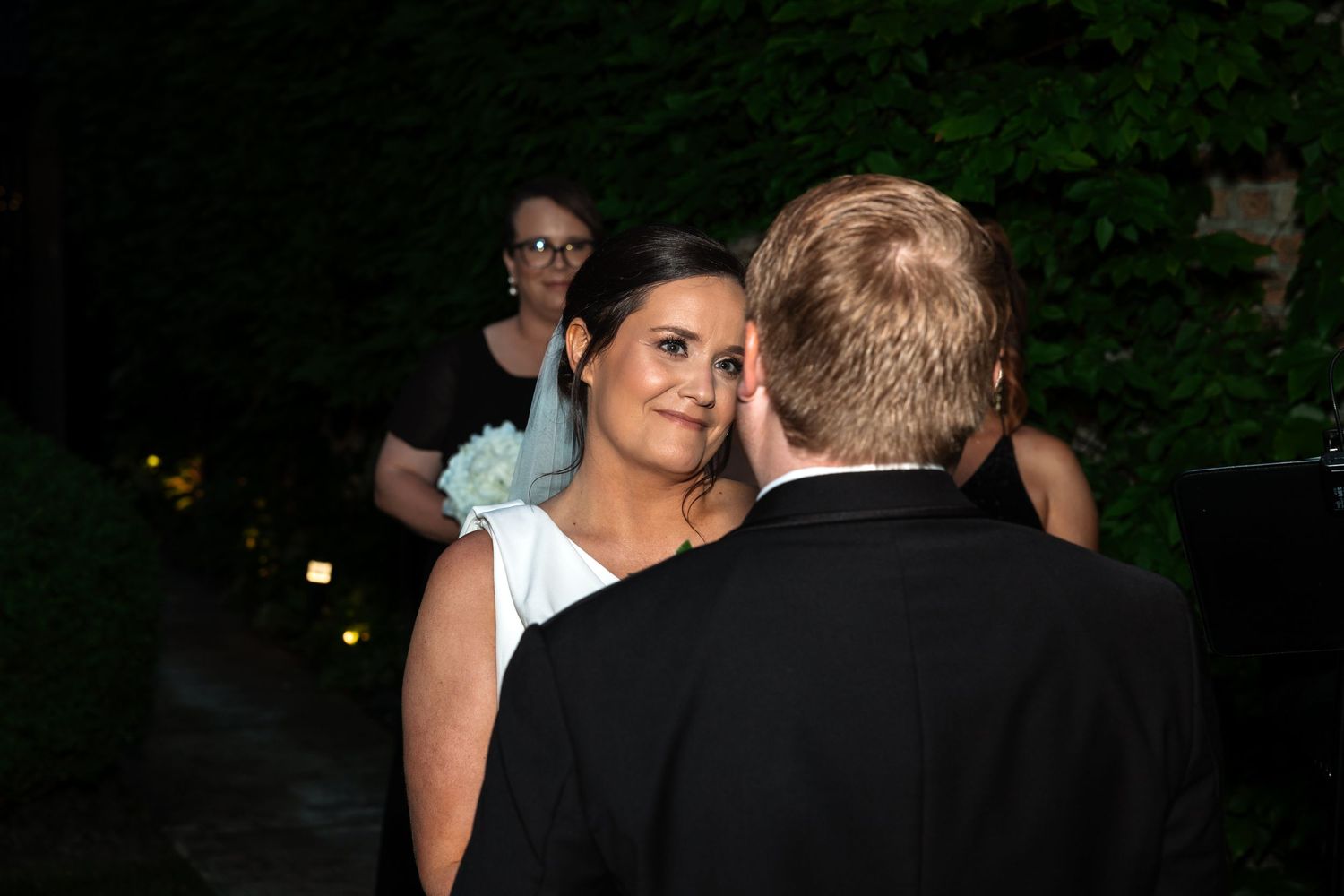 Bride in white dress smiles lovingly at groom during evening outdoor wedding ceremony with guests behind.