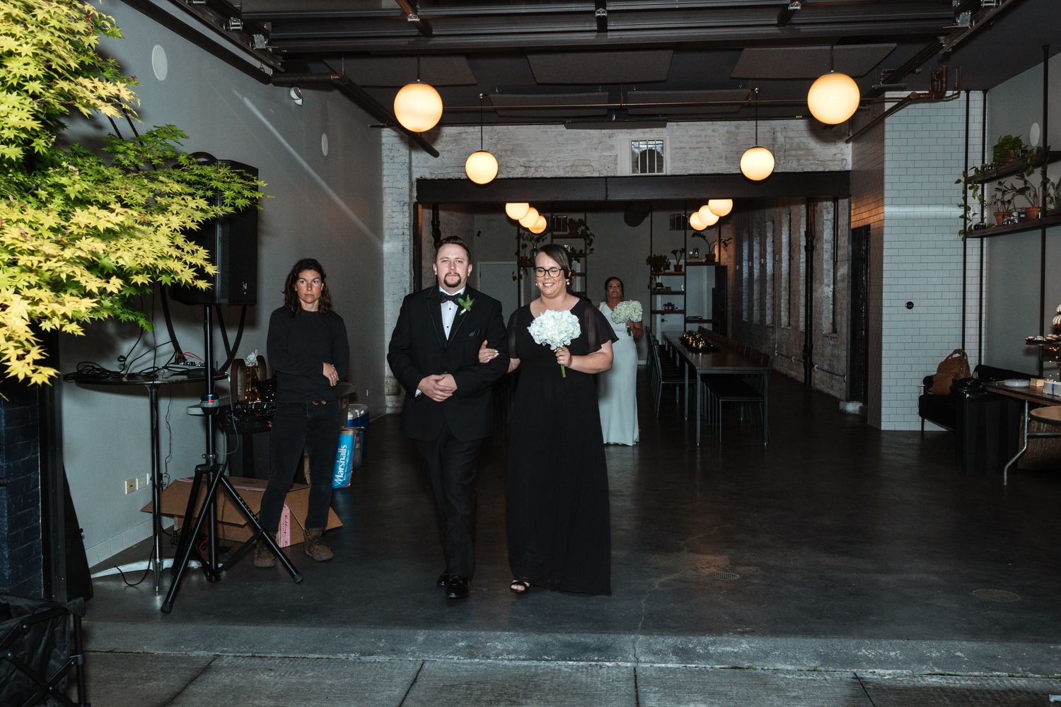 A couple walks down the aisle at an indoor wedding venue with globe pendant lights and lush greenery.