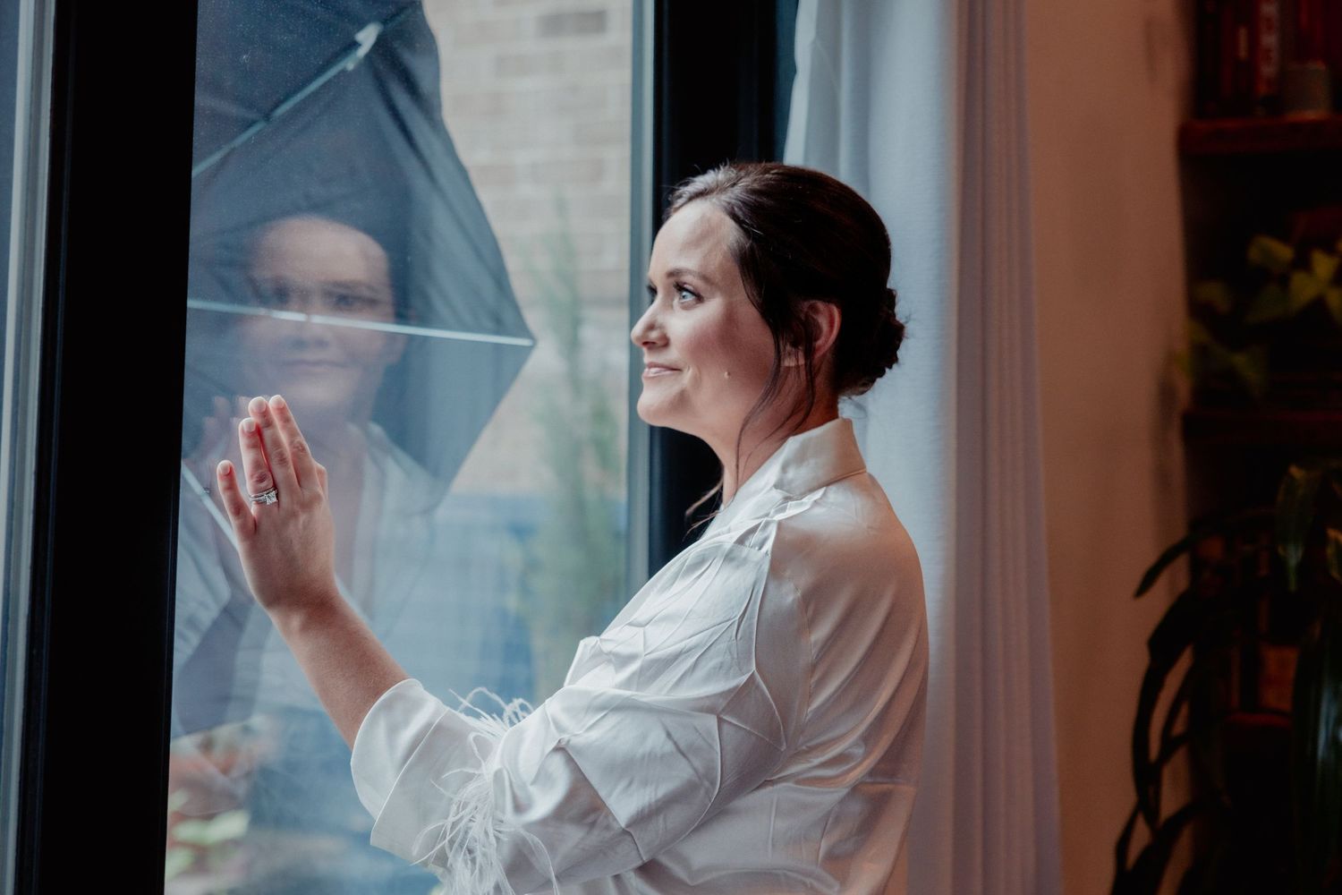 Bride in white robe gazing out window with her reflection visible in the glass.