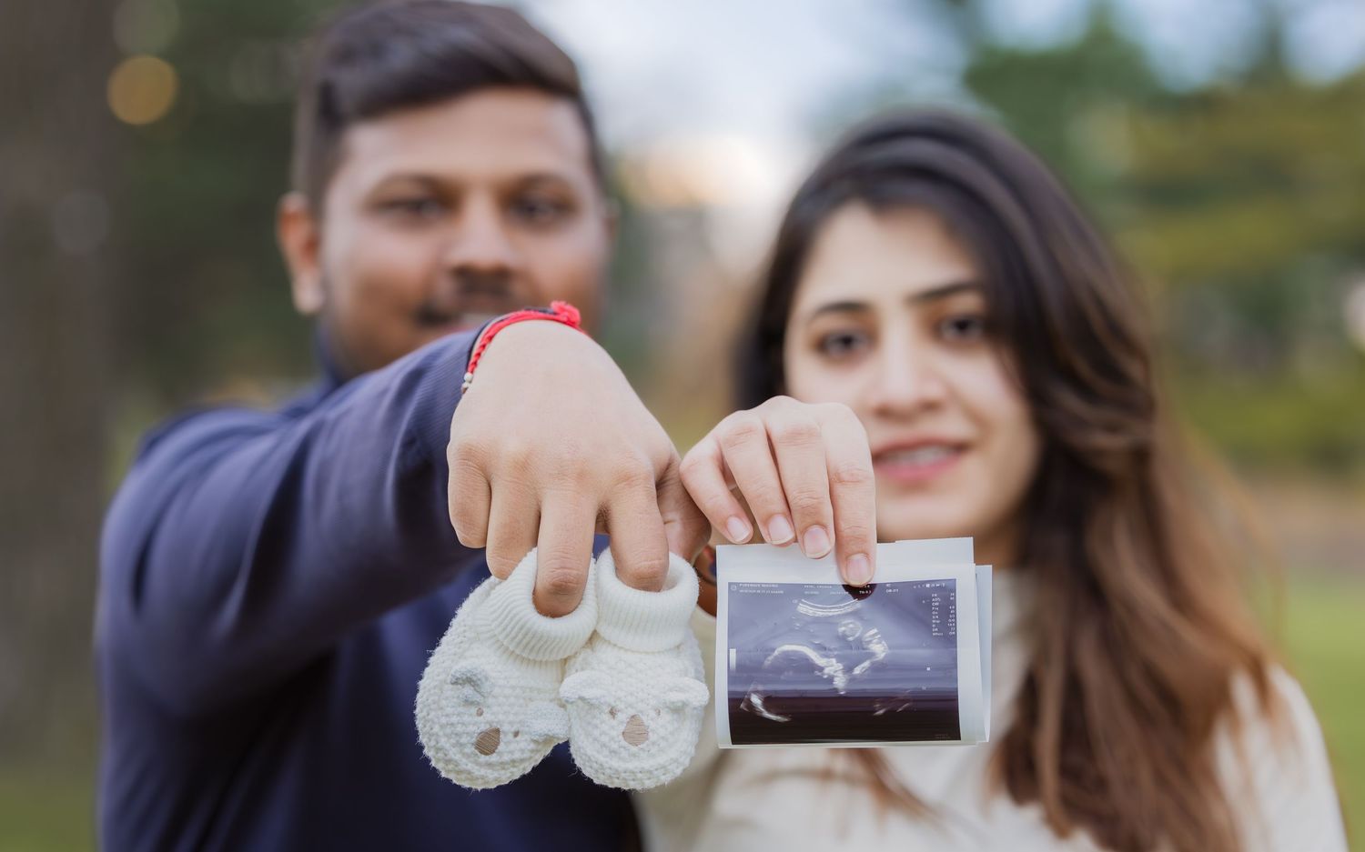Couple displaying baby booties and ultrasound scan during outdoor maternity announcement.