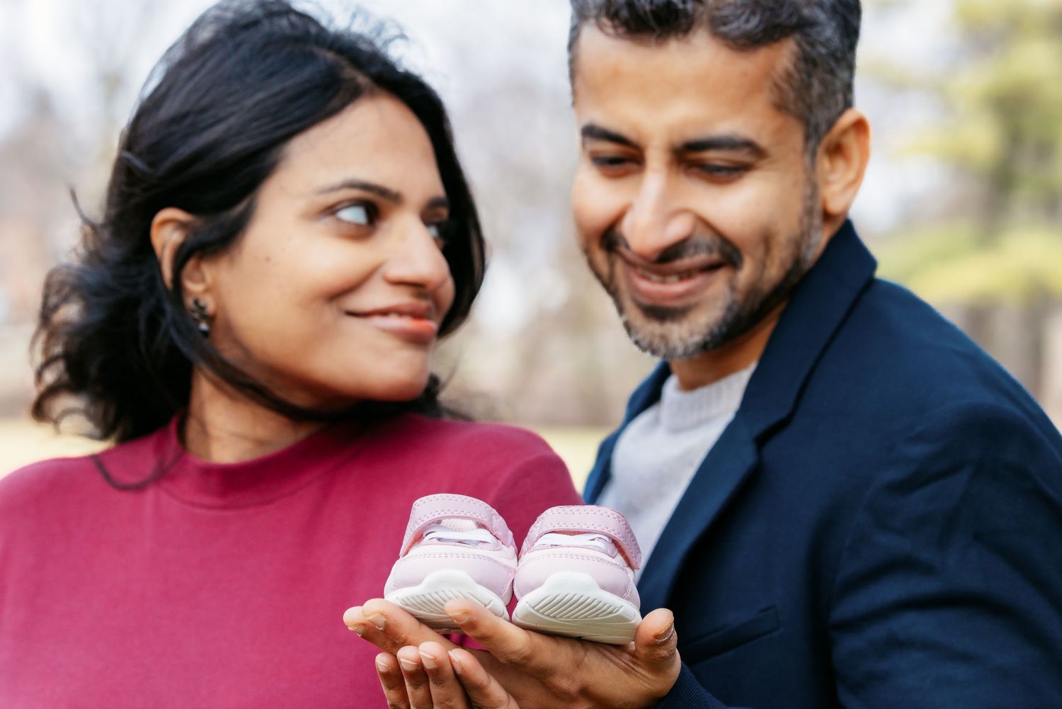 Smiling expecting couple holding tiny pink baby shoes together in an outdoor setting.