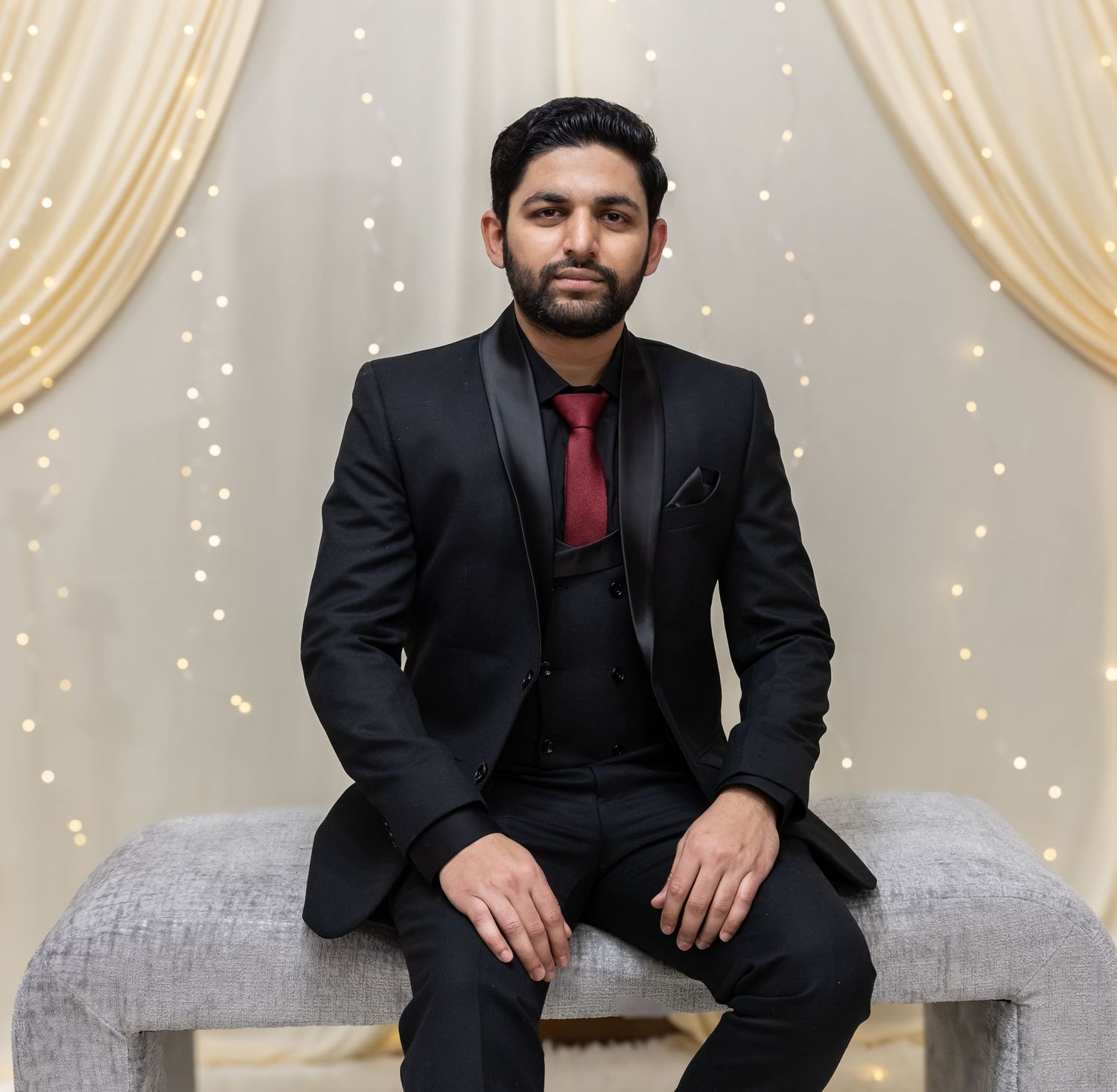 A man in a black tuxedo sits elegantly on a bench at a wedding venue with twinkling lights.