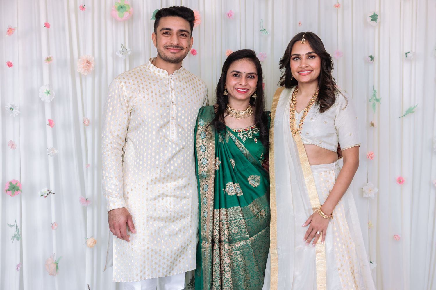 Trio posing at Indian celebration, woman in green saree flanked by couple in white outfits.