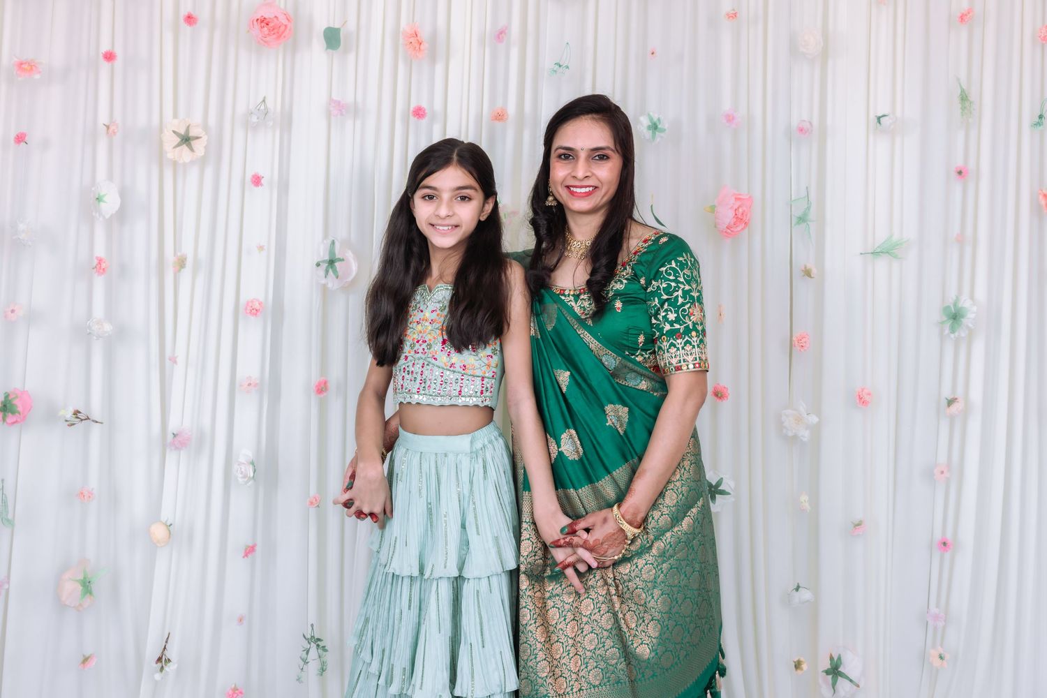 Mother and daughter in traditional Indian green outfits smile together at festive event with floral backdrop.