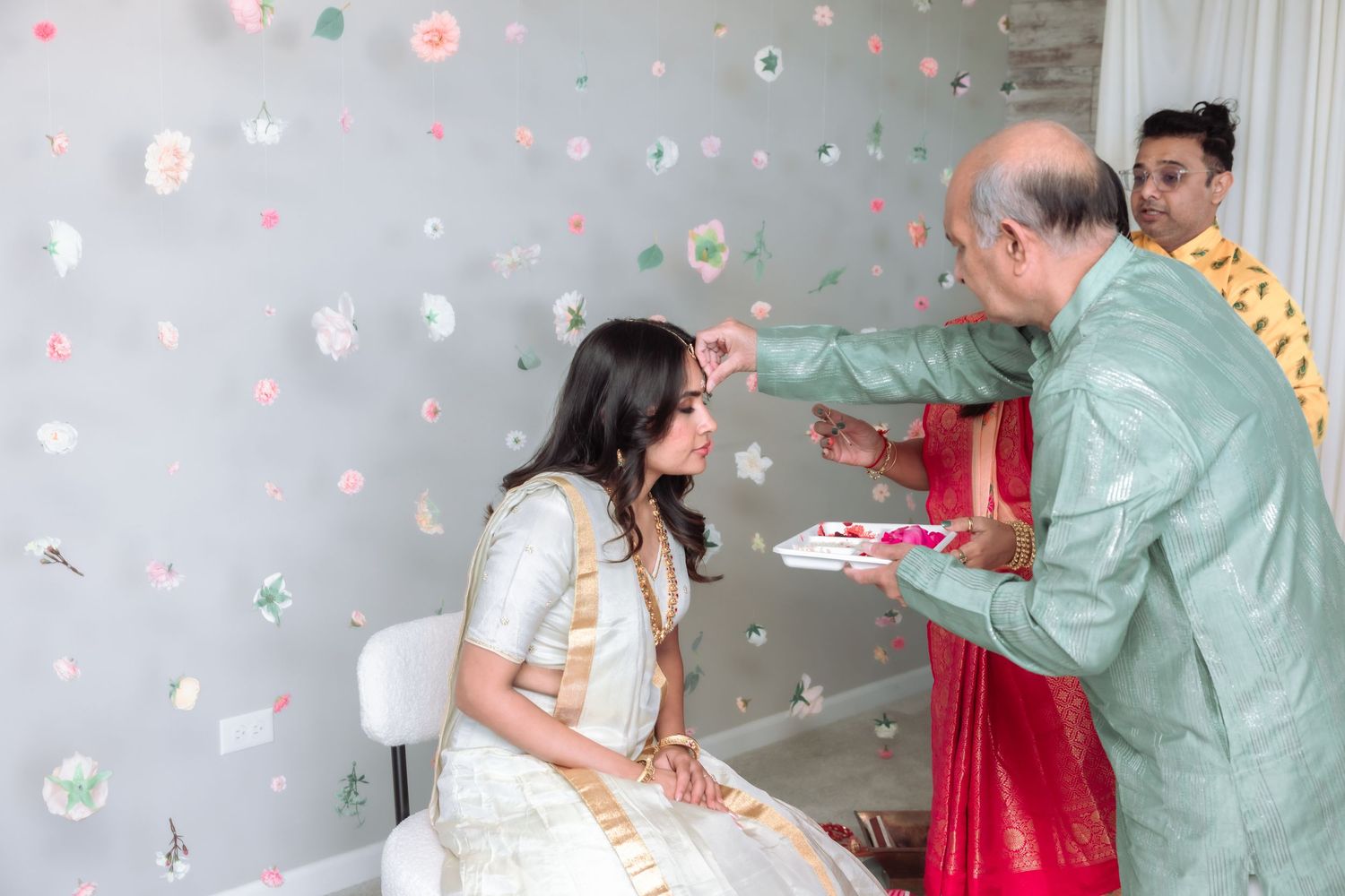 Elder man applying tilak to young woman in white saree during Indian wedding ceremony with floral backdrop.
