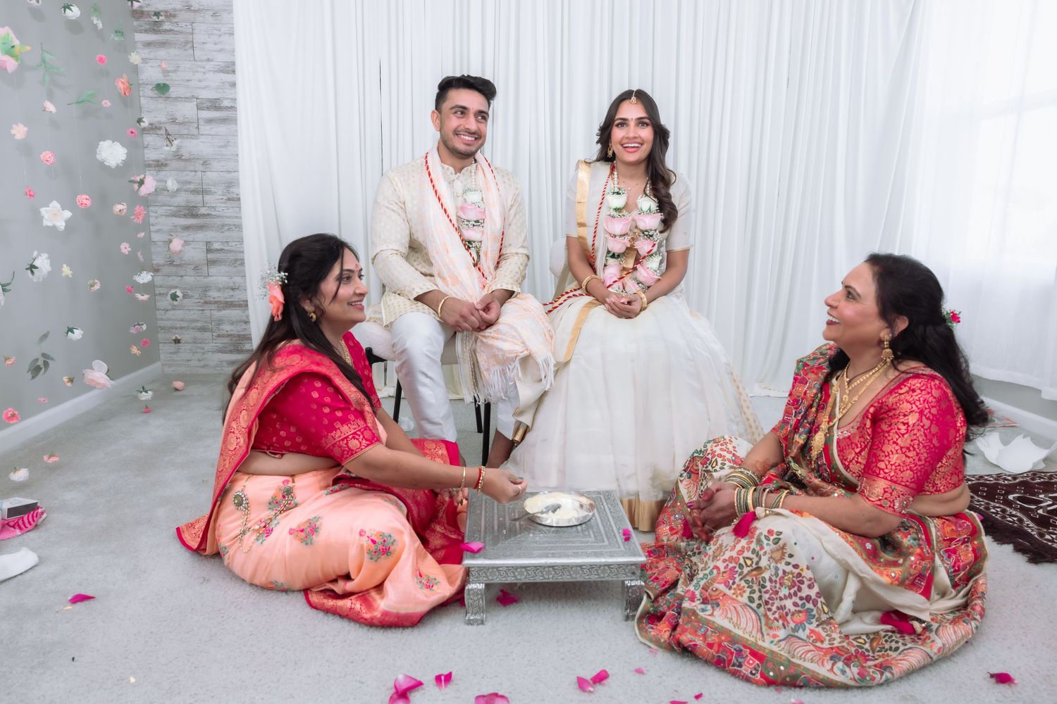 Indian wedding couple seated with two women in red sarees performing traditional ceremony ritual.