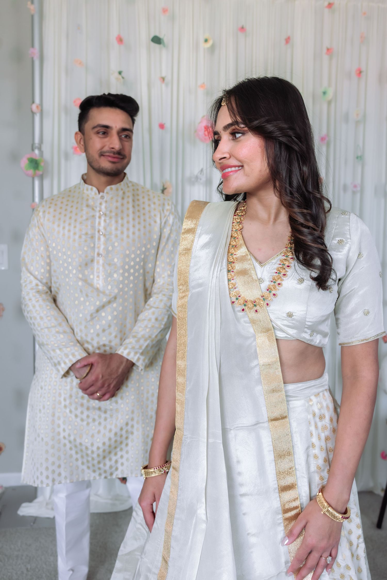 Indian couple in matching white and gold traditional outfits posing at their elegant engagement ceremony.