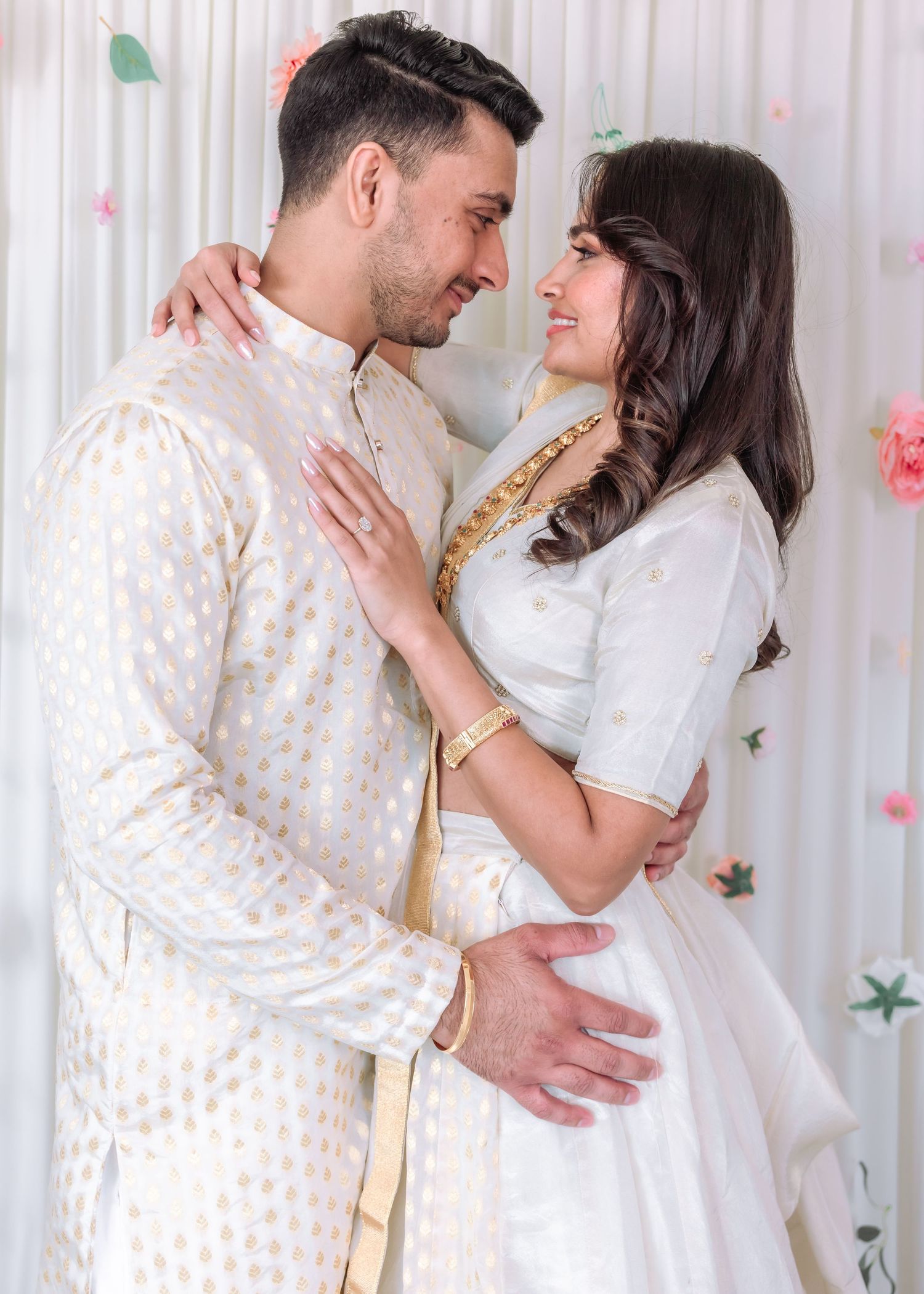 Indian couple in white and gold traditional attire share a romantic embrace at a floral ceremony.
