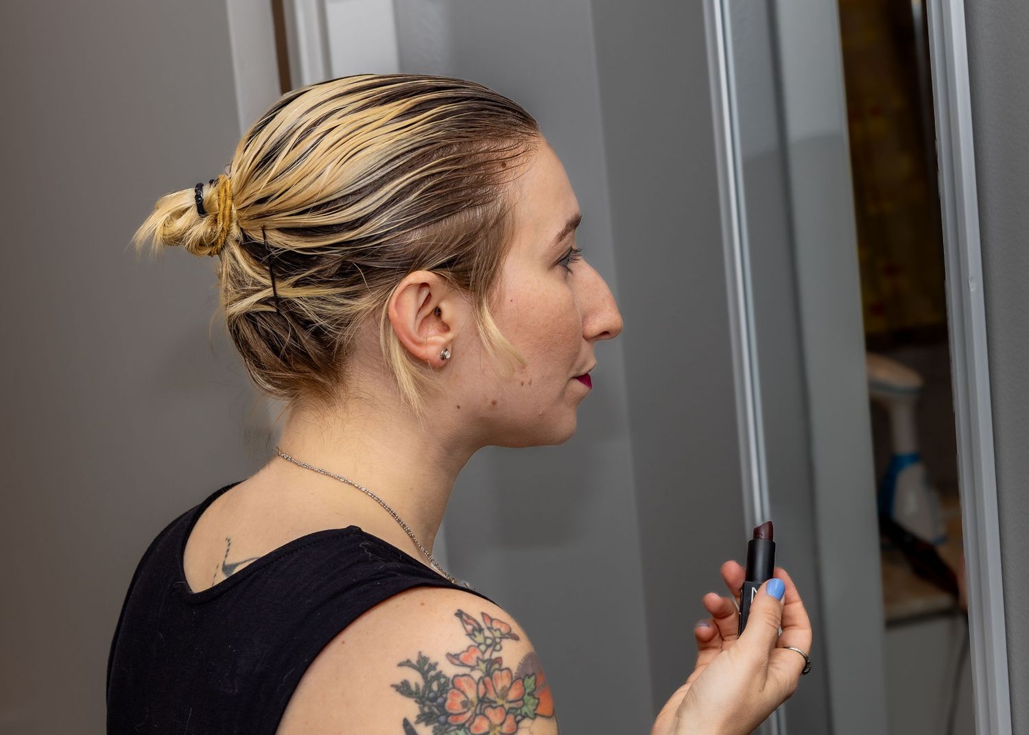 Young blonde woman with floral shoulder tattoo and hair in bun looking at mirror holding makeup tool.