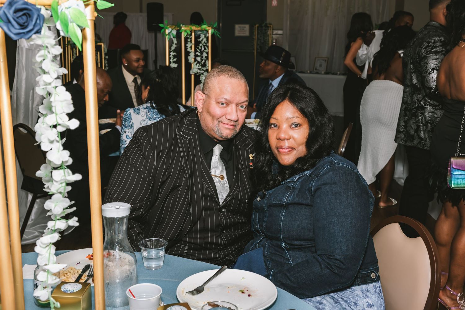 A couple in formal attire smiles together at a decorated wedding reception table with floral arrangements.