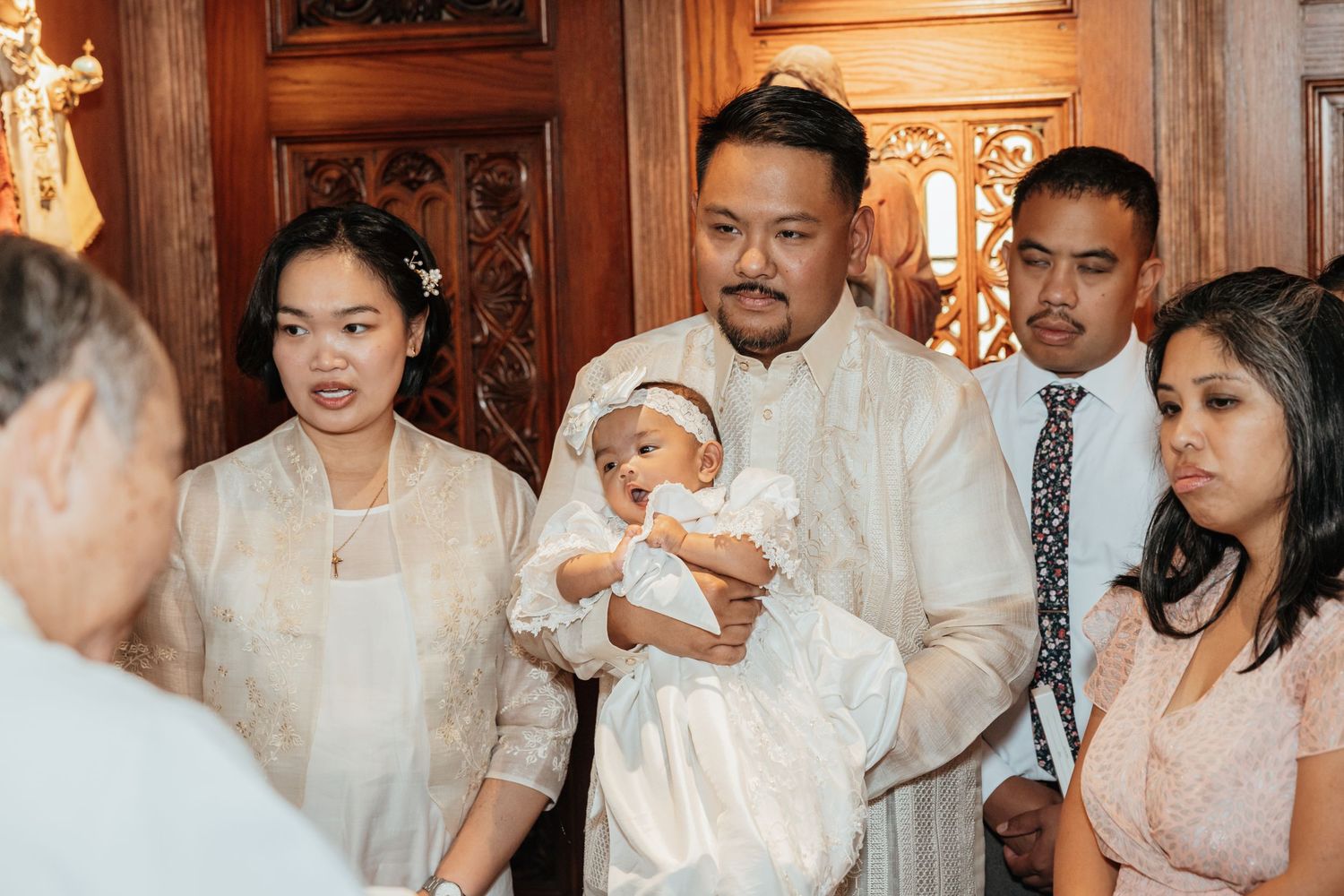 Filipino family in white barong attire gather around a priest during a Catholic baby baptism ceremony.