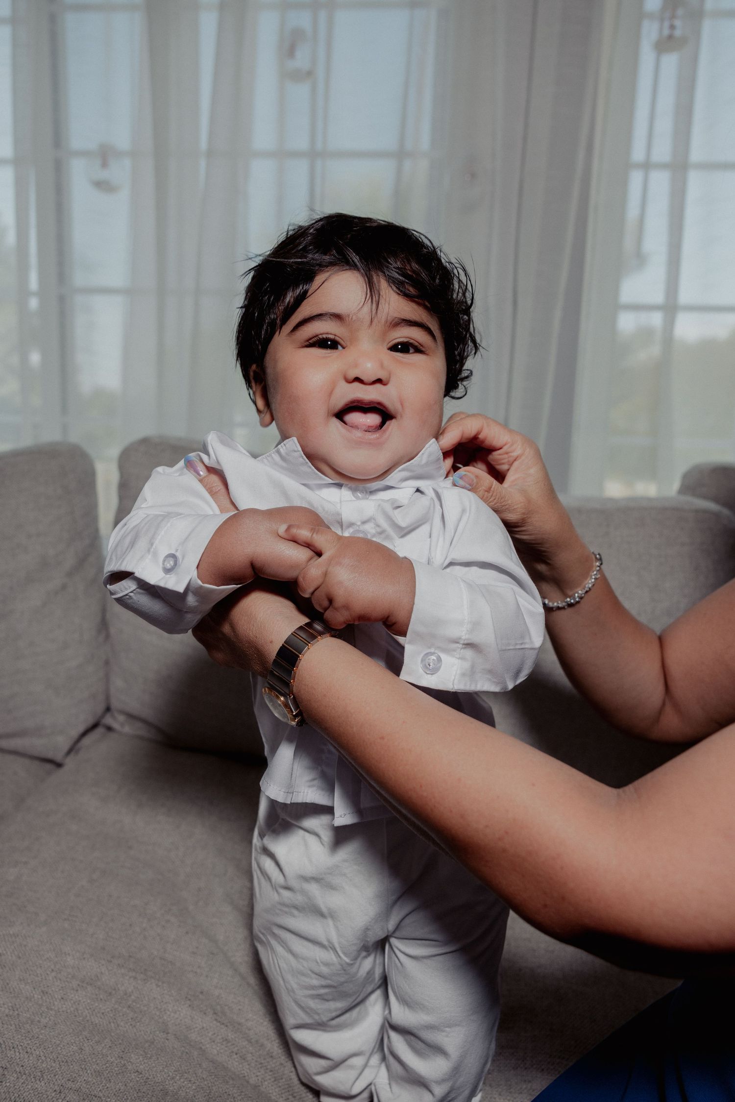 Happy baby in white christening outfit being held up on sofa, smiling and laughing.